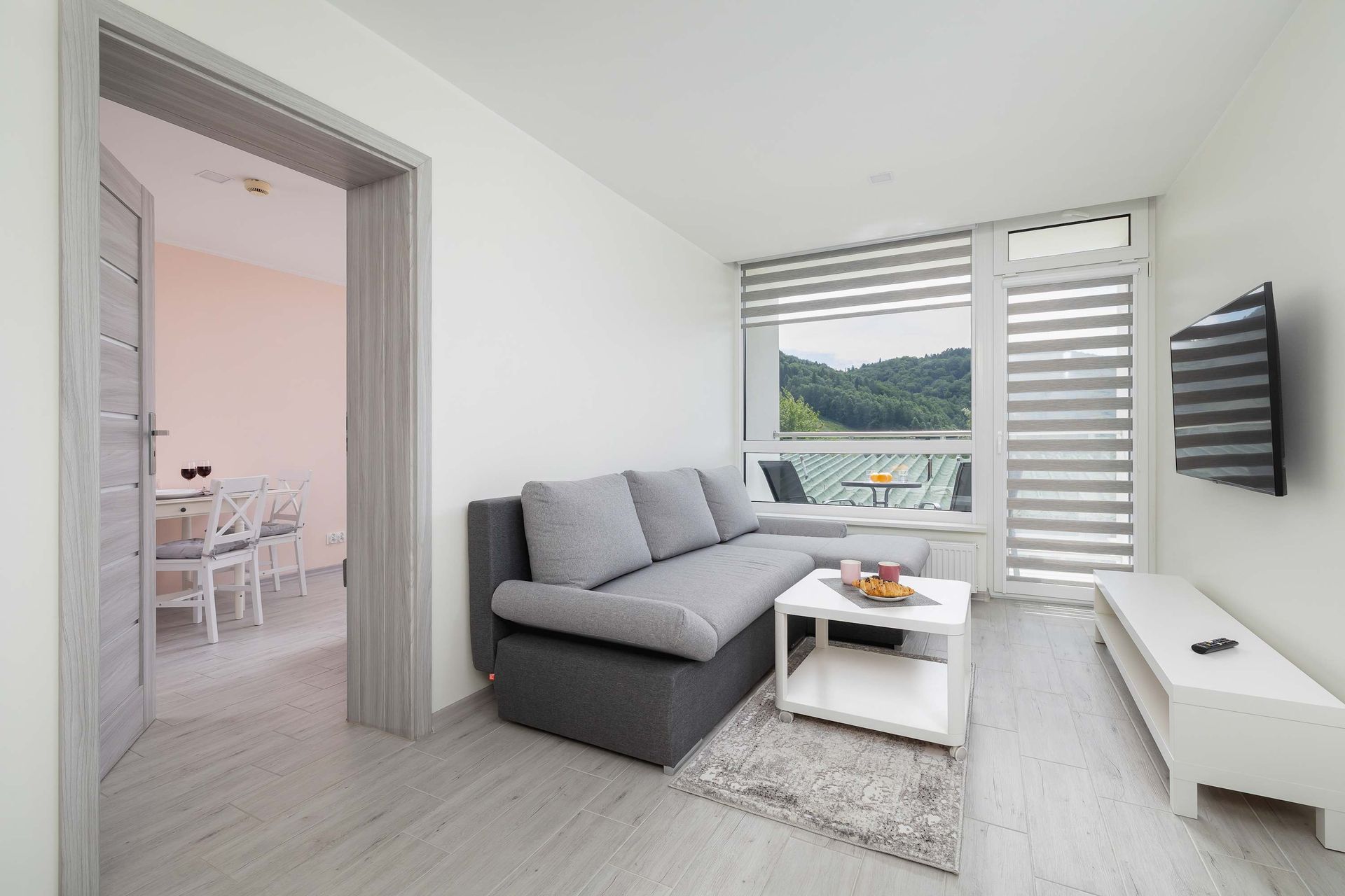 Przytulny salon z szarą sofą, białym stolikiem kawowym i dużym oknem z widokiem na las.