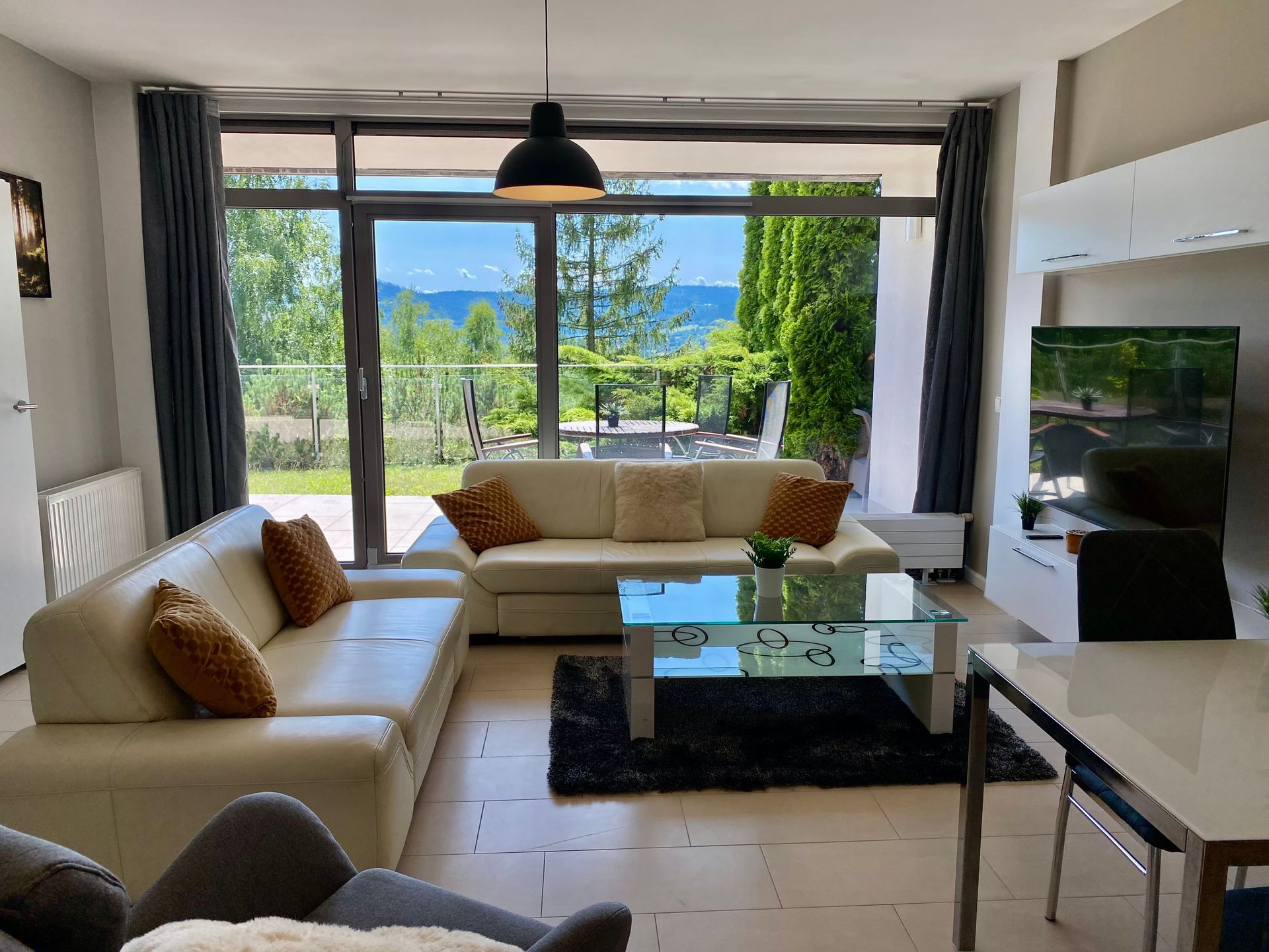 Salon z białymi sofami, szklanym stolikiem kawowym i dużym oknem z widokiem na taras i góry.