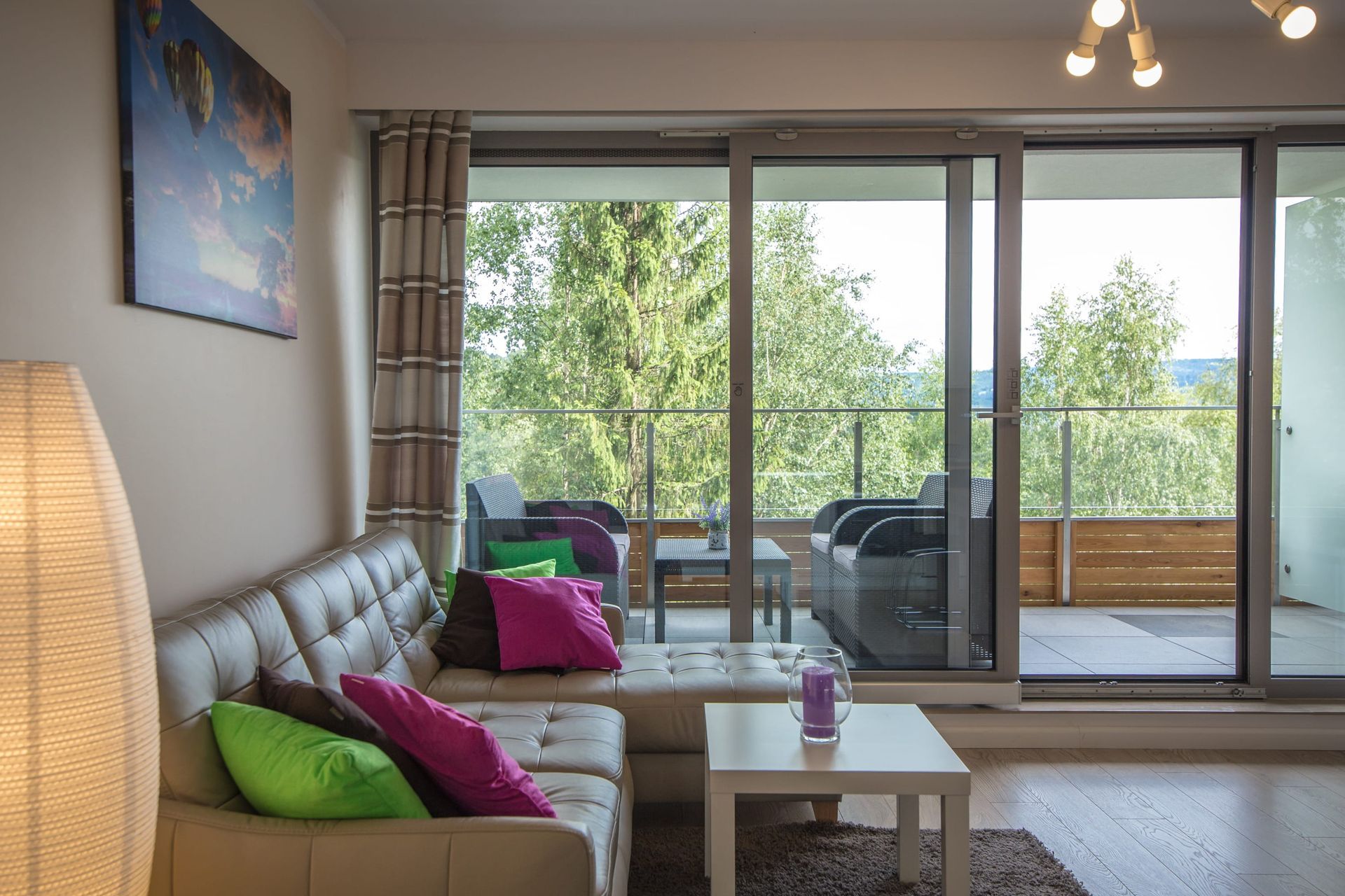 Salon z jasną sofą, kolorowymi poduszkami i szklanymi drzwiami przesuwnymi prowadzącymi na balkon z widokiem na góry i drzewa.