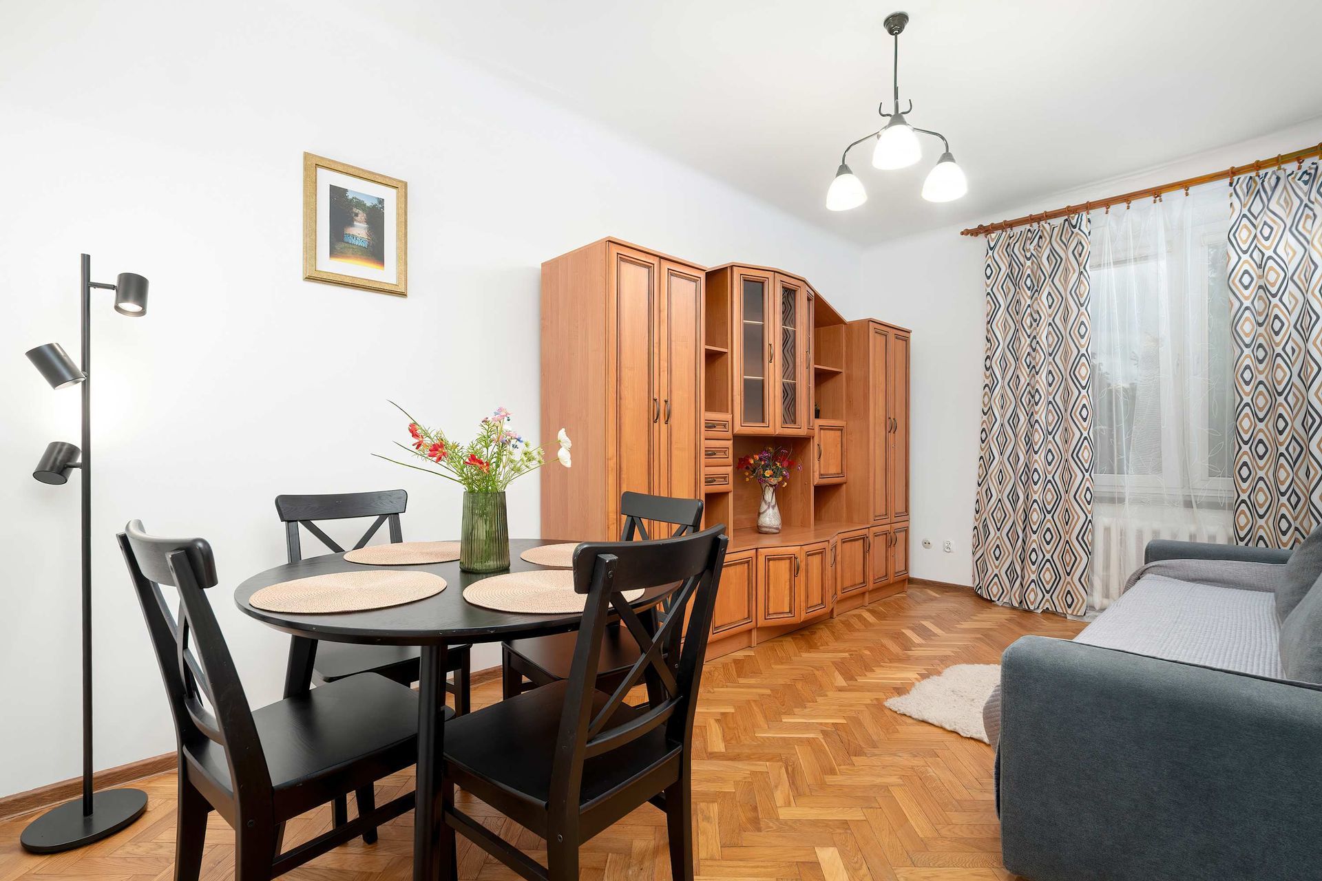 Przytulny salon z jadalnią z czarnym stołem, drewnianą szafką, szarą sofą i wzorzystymi zasłonami