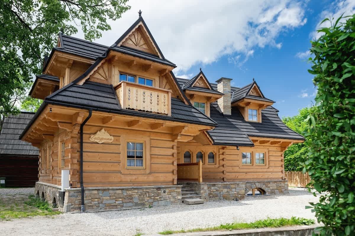 Drewniany dom w stylu chalet z oknami dachowymi i kamienną podstawą, otoczony drzewami pod błękitnym niebem