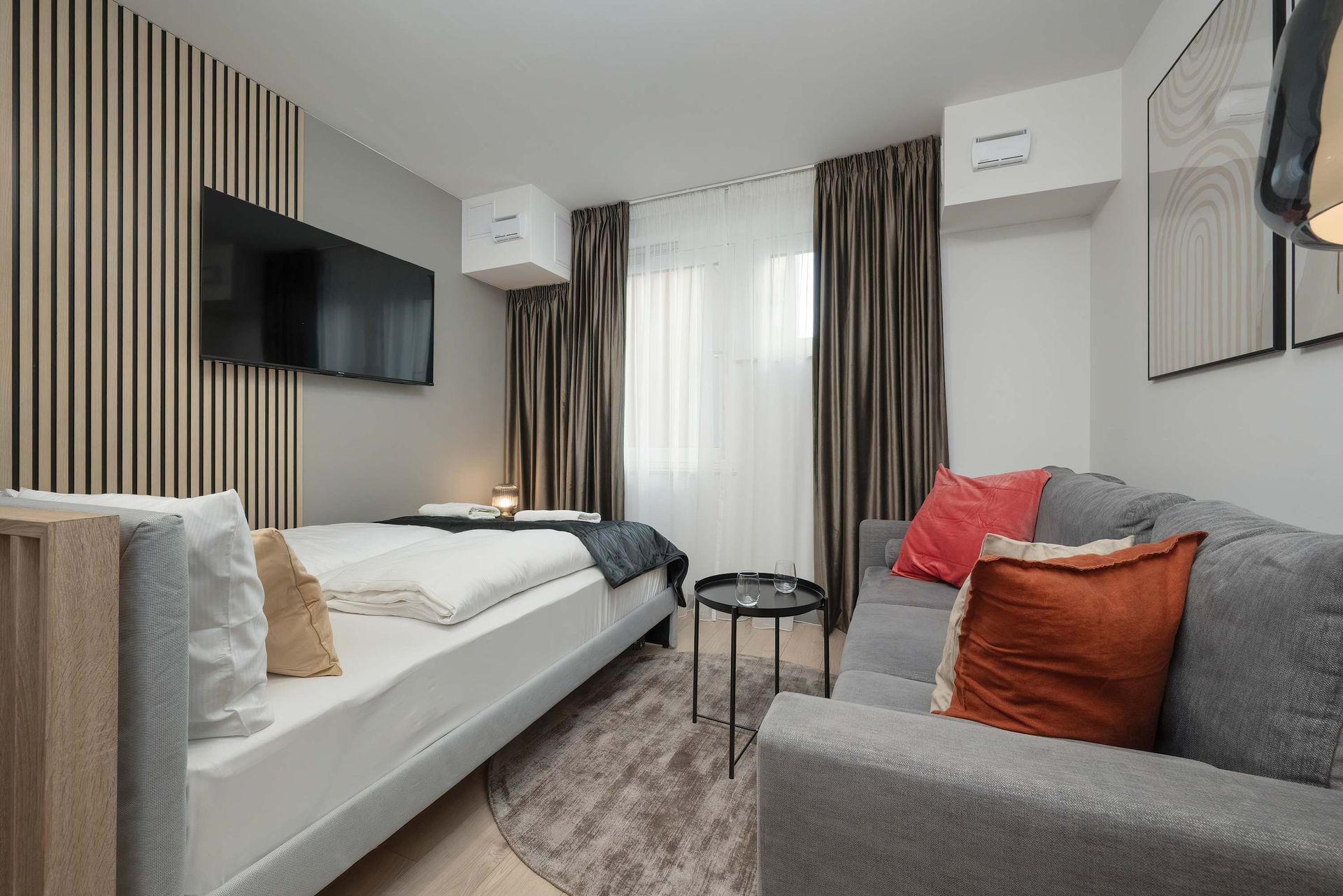 Mała sypialnia w mieszkaniu z łóżkiem, sofą, telewizorem i dekoracyjnymi panelami ściennymi.