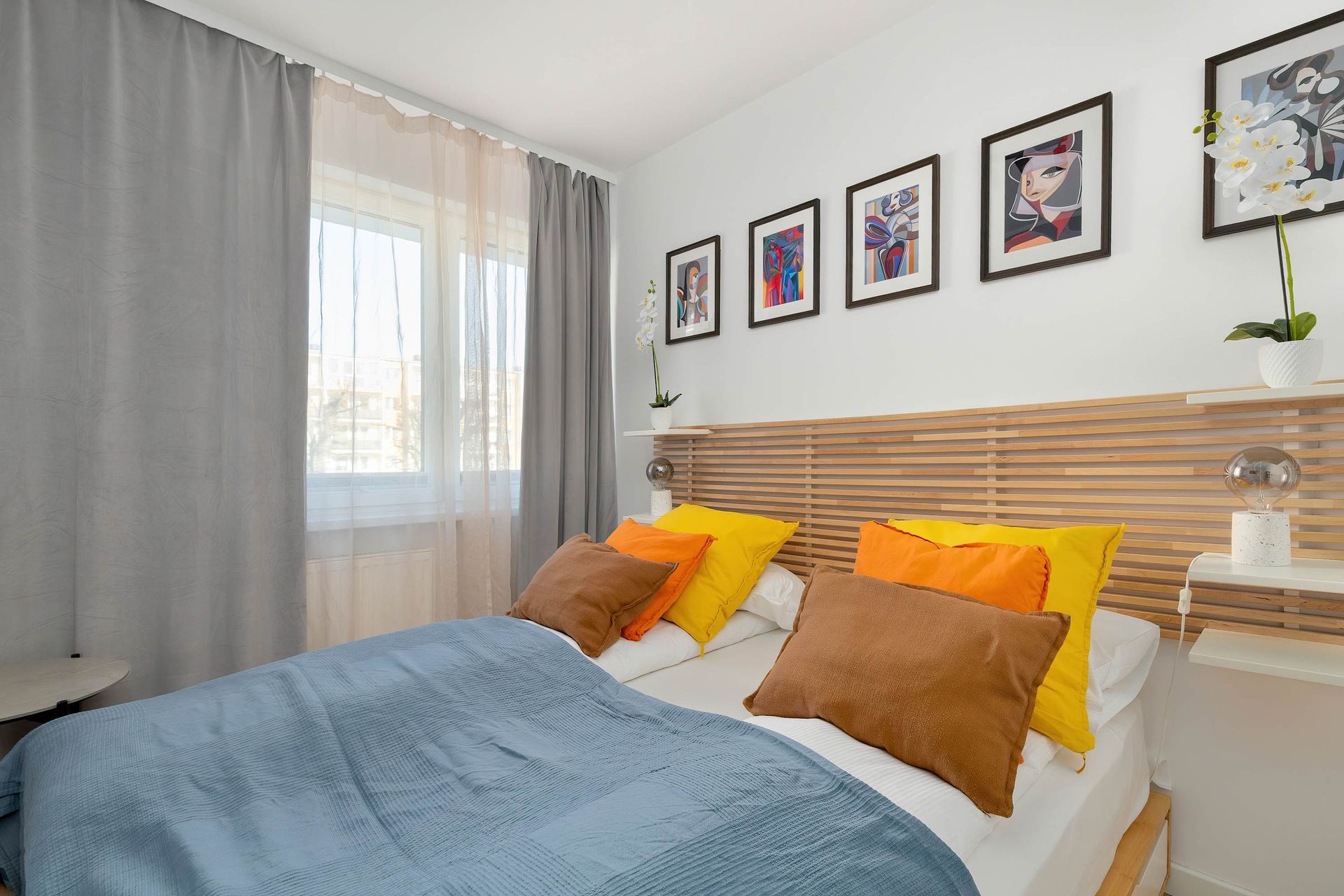 Przytulna sypialnia z łóżkiem z niebieskim narzutem, pomarańczowymi i żółtymi poduszkami, zagłówkiem z drewnianych listew i grafiką ścienną.