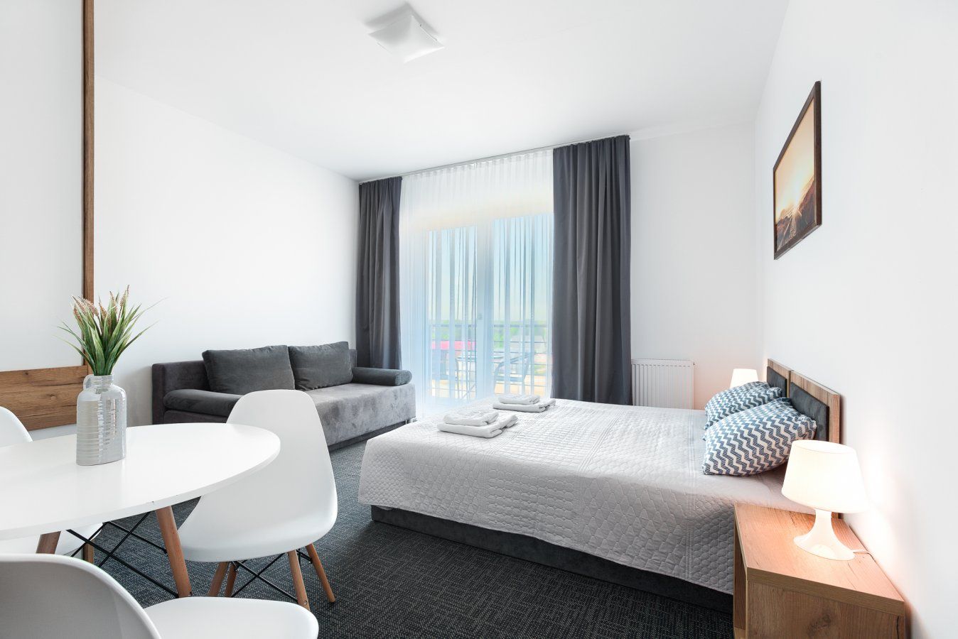 Kawalerka z łóżkiem, sofą, stołem jadalnym i wyjściem na balkon. Białe ściany, szare zasłony i dywan.