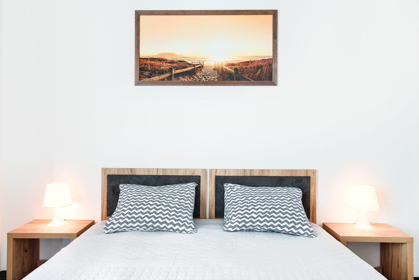 Sypialnia z łóżkiem, dwiema szafkami nocnymi, lampami i obrazem przedstawiającym zachód słońca.