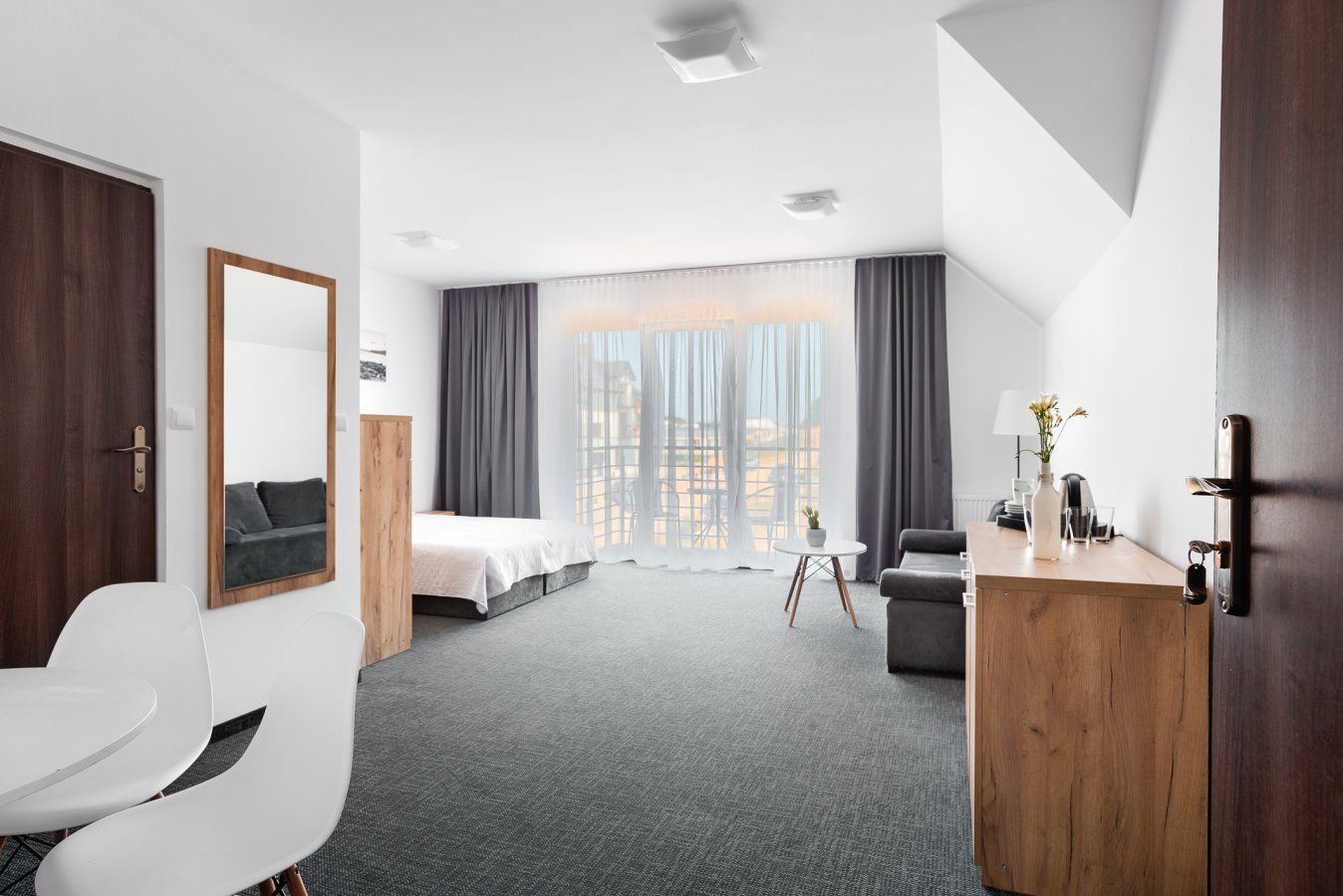 Wnętrze pokoju hotelowego z łóżkiem, meblami, dużym oknem i otwartymi drzwiami.