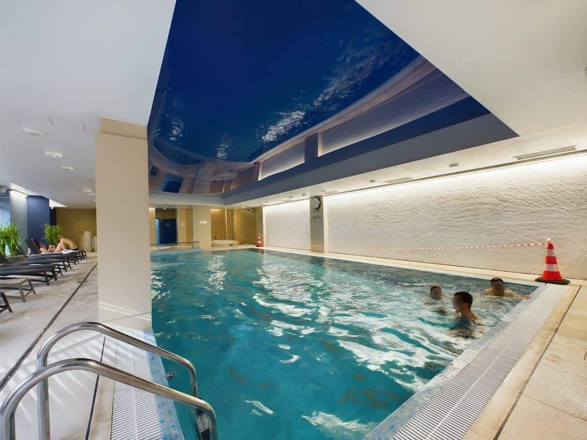 Kryty basen z niebieskim oświetleniem, kafelkową ścianą, drabinką, leżakami i pływającymi ludźmi.