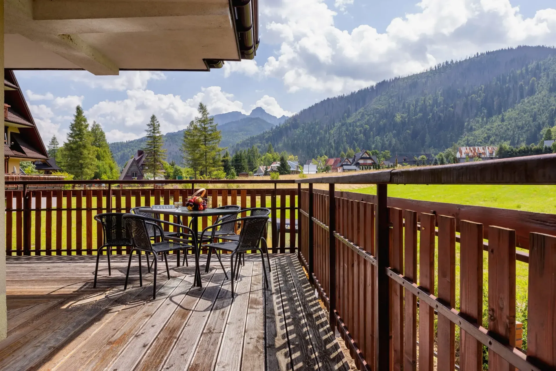 Balkon ze stołem i krzesłami, z widokiem na zielone pole i pasmo górskie w słoneczny dzień.