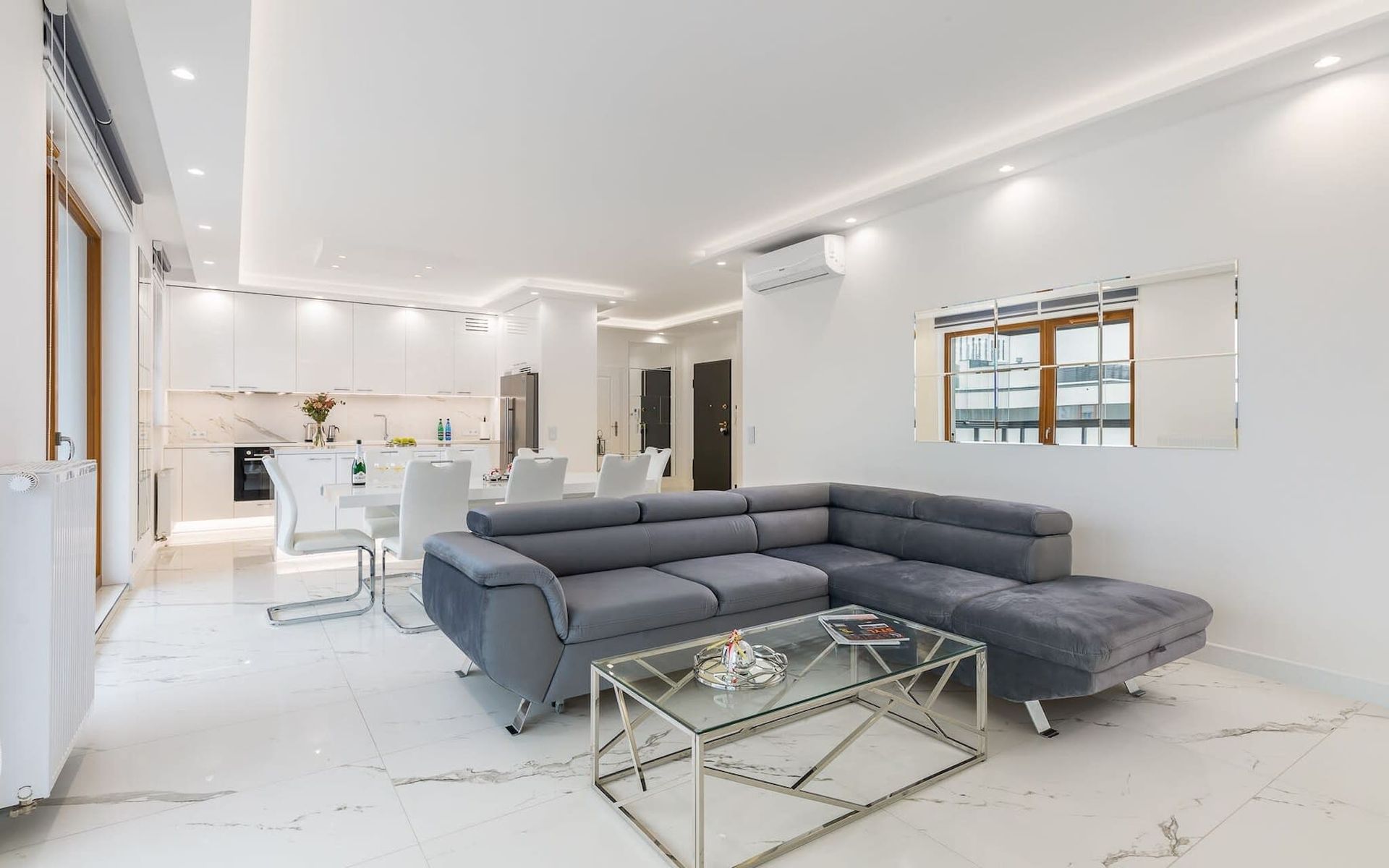 Nowoczesny salon z szarą narożną sofą, otwarty na białą kuchnię i jadalnię.