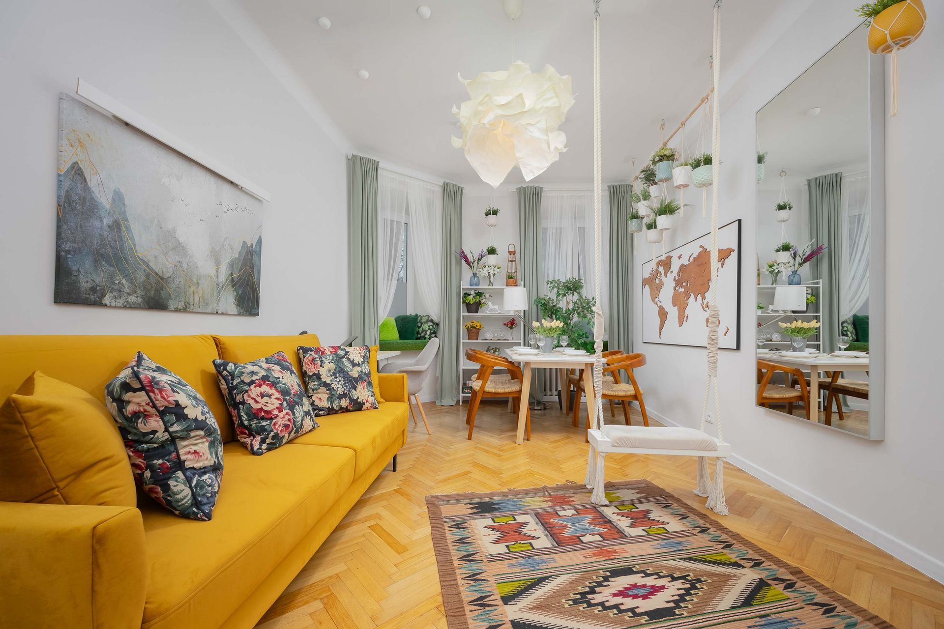 Jasny salon z żółtą sofą, wzorzystym dywanem, stołem jadalnym i grafiką ścienną, z lustrzanymi ścianami i ciepłą drewnianą podłogą