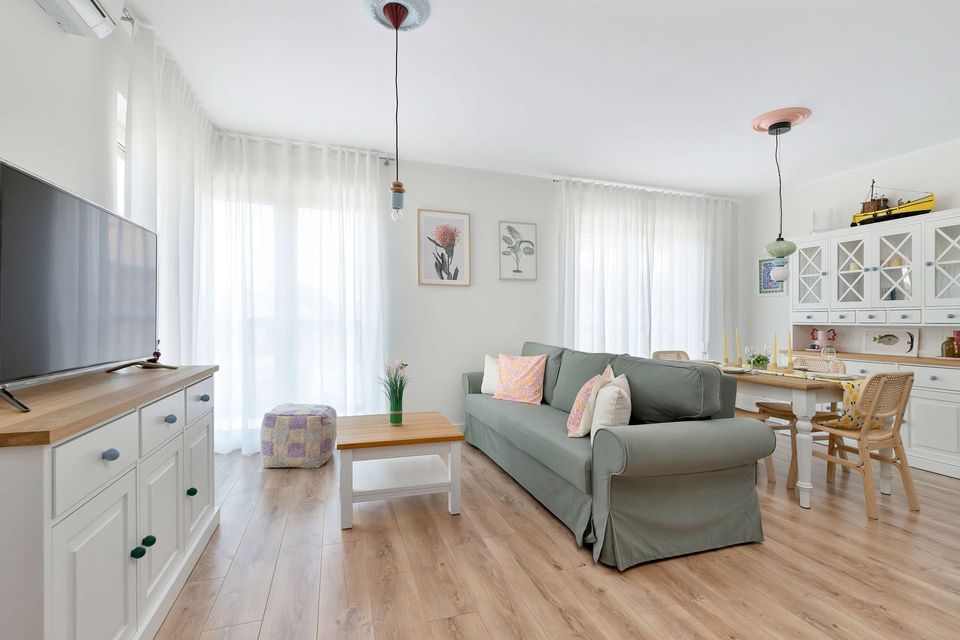 Jasny salon z szarą sofą, drewnianym stolikiem kawowym, telewizorem, jadalnią i cienkimi, białymi zasłonami