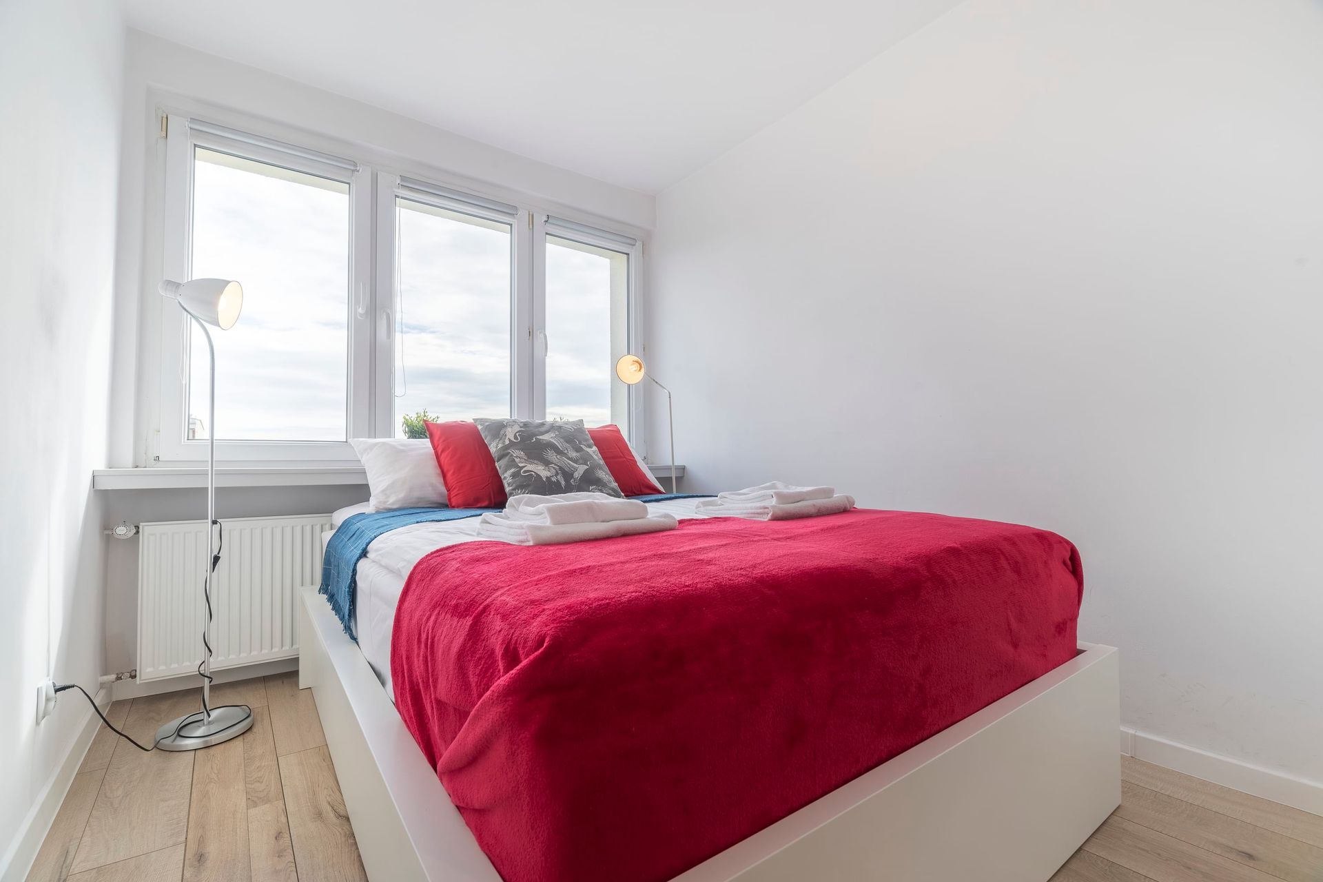 Jasna sypialnia z białym łóżkiem, jaskrawoczerwonym kocem, dekoracyjnymi poduszkami i stojącą lampą przy oknie.