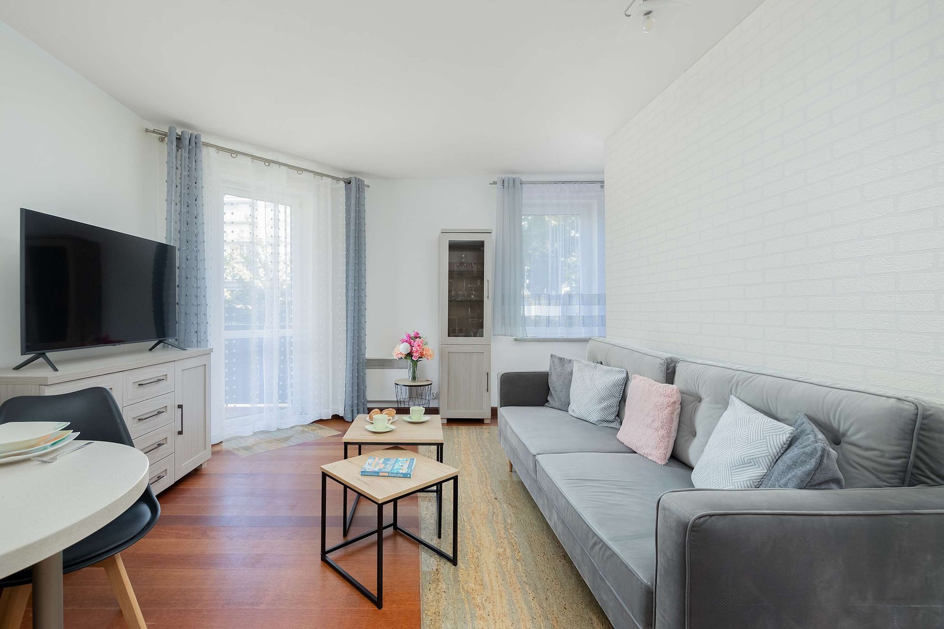 Jasny, nowoczesny salon z szarą sofą, drewnianymi stolikami kawowymi, telewizorem na kredensie i białymi ceglanymi ścianami.