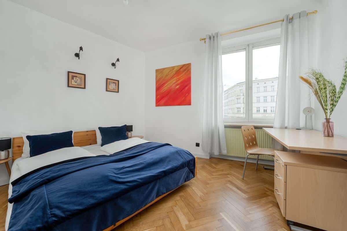 Jasna sypialnia z niebieskim łóżkiem, drewnianym biurkiem, białymi zasłonami i kolorowymi dekoracjami ściennymi