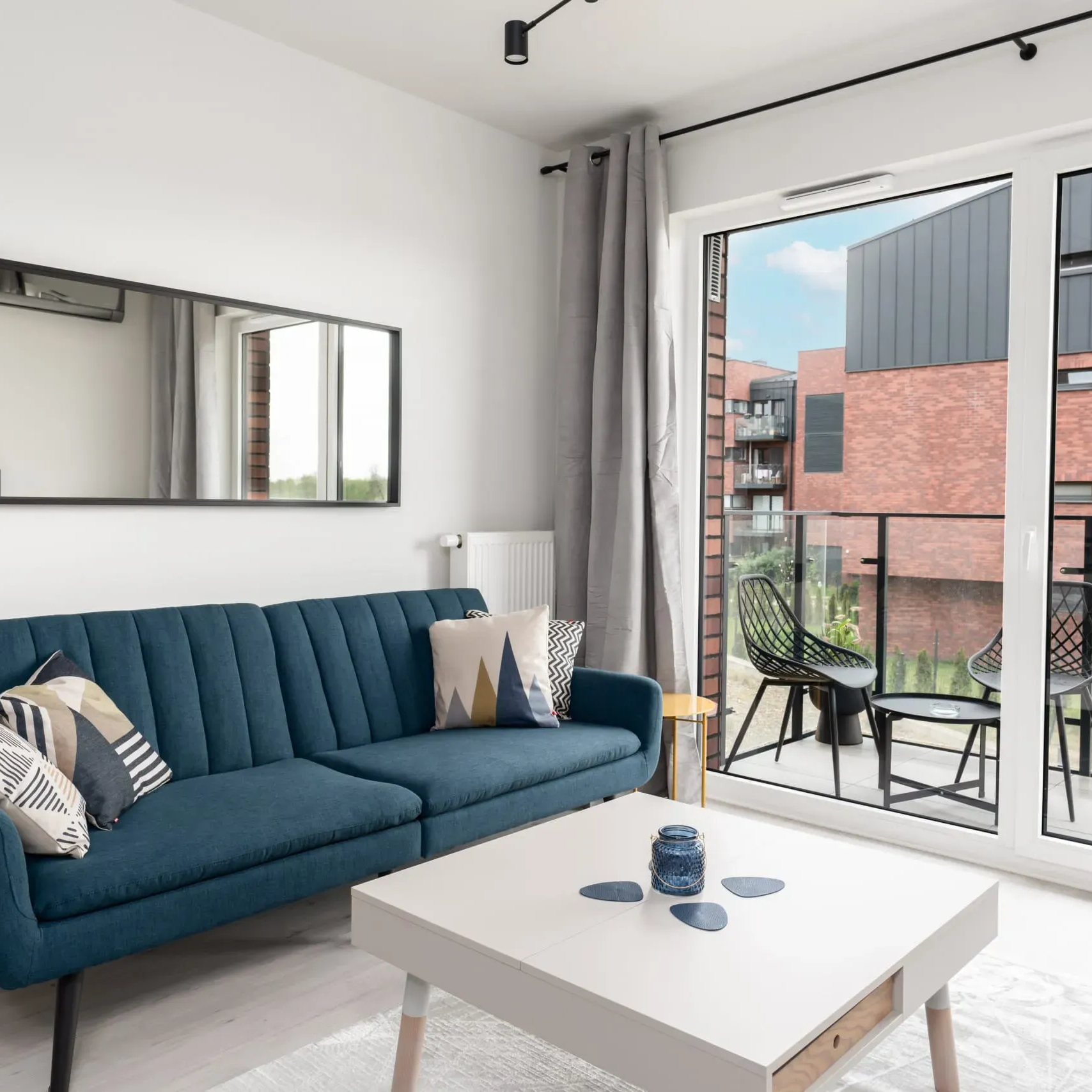 Salon z niebieską sofą, białym stolikiem kawowym i balkonem z widokiem na ceglany budynek.