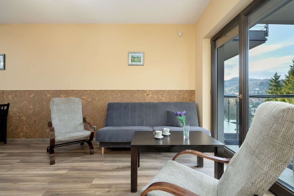 Nowoczesny salon z beżowymi ścianami, szarą sofą, fotelami, stolikiem kawowym i dużymi przesuwanymi drzwiami szklanymi.