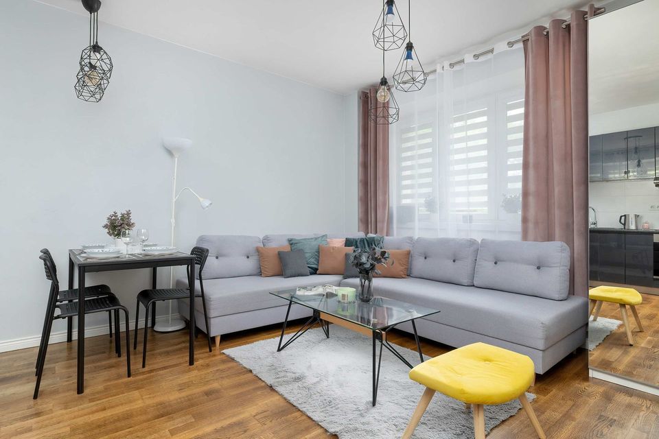 Nowoczesny salon z szarą sofą modułową, szklanym stolikiem kawowym, żółtym stołkiem i małą jadalnią przy oknie