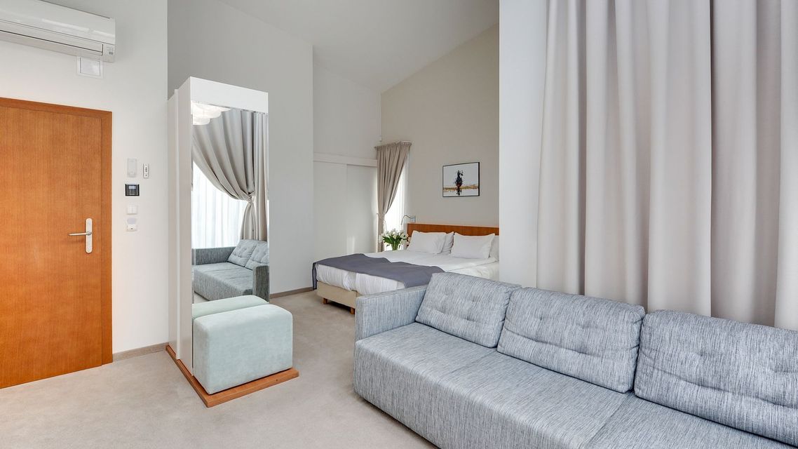 Nowoczesny pokój hotelowy z szarą sofą, kącikiem sypialnym, białymi zasłonami i drewnianymi drzwiami