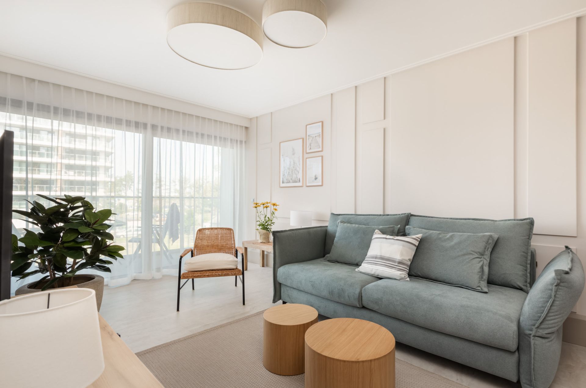 Salon z niebieską sofą, drewnianymi stolikami nocnymi, rattanowym krzesłem, dużym oknem i rośliną.