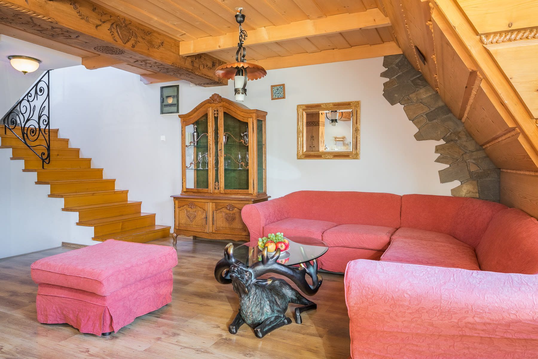 Rustykalny salon z czerwoną narożną sofą, pasującą do niej pufą, drewnianą szafką i schodami prowadzącymi na piętro.