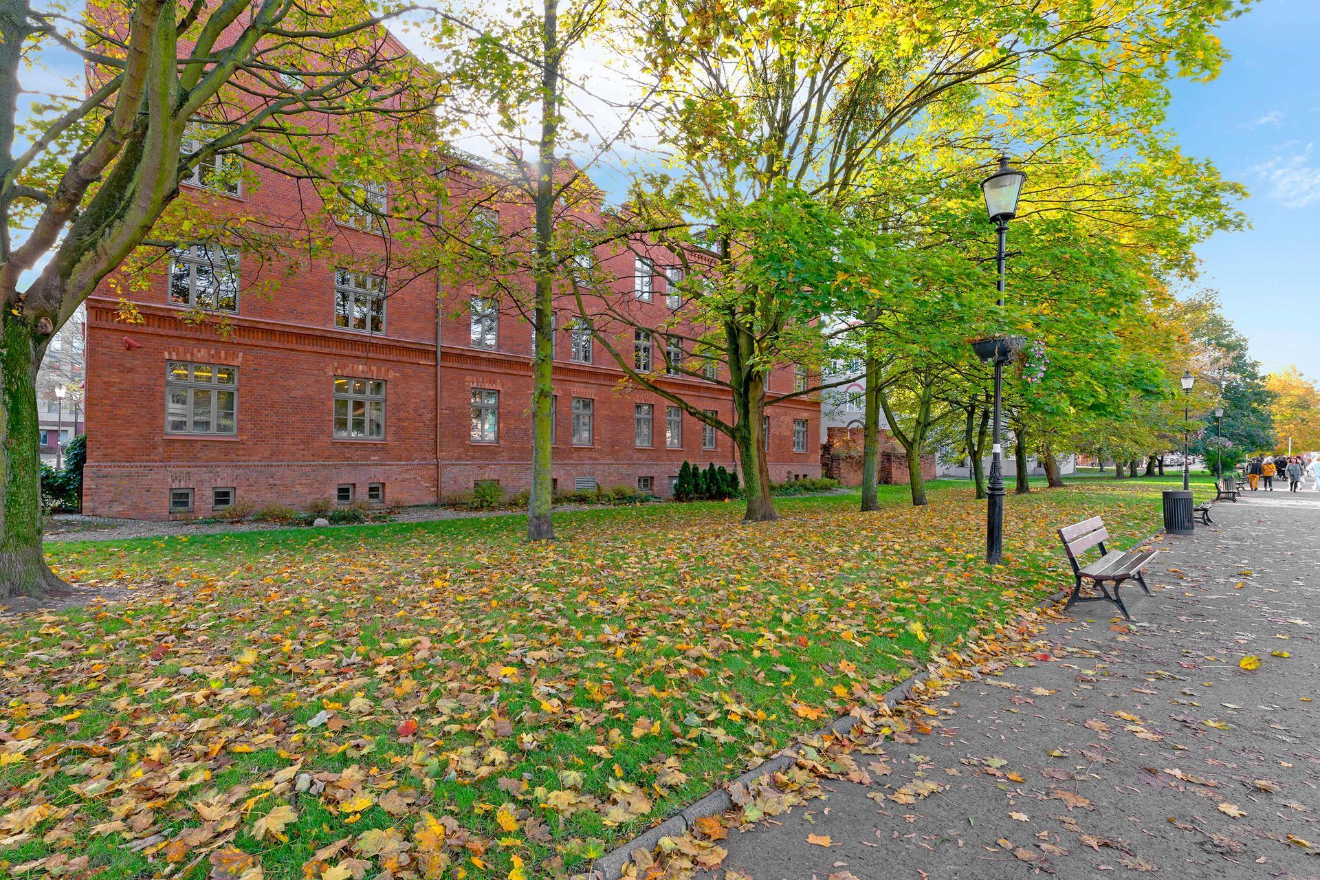 Czerwony ceglany budynek z drzewami i opadłymi liśćmi w otoczeniu parku; po prawej stronie alejka z ławkami.