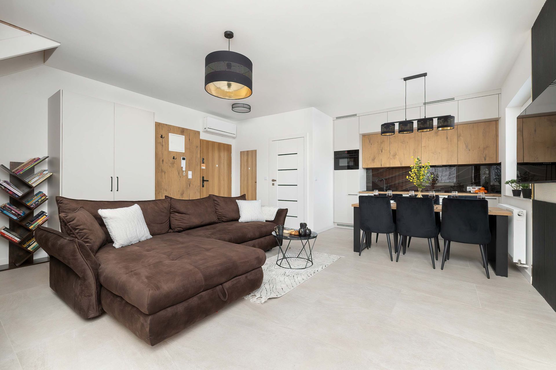 Jasny, otwarty salon połączony z kuchnią z brązową sofą modułową, czarnymi krzesłami do jadalni i szafkami z jasnego drewna.