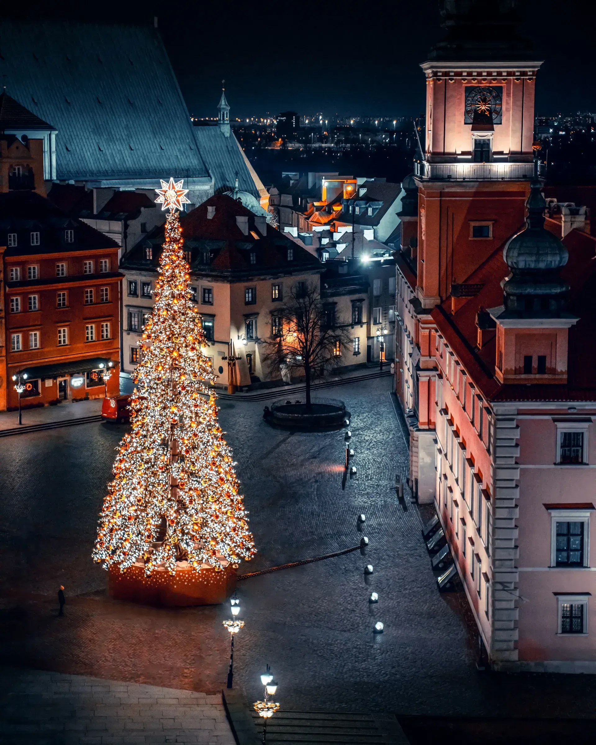 Oświetlona choinka na brukowanym placu w centrum Warszawy, otoczona budynkami w nocy.