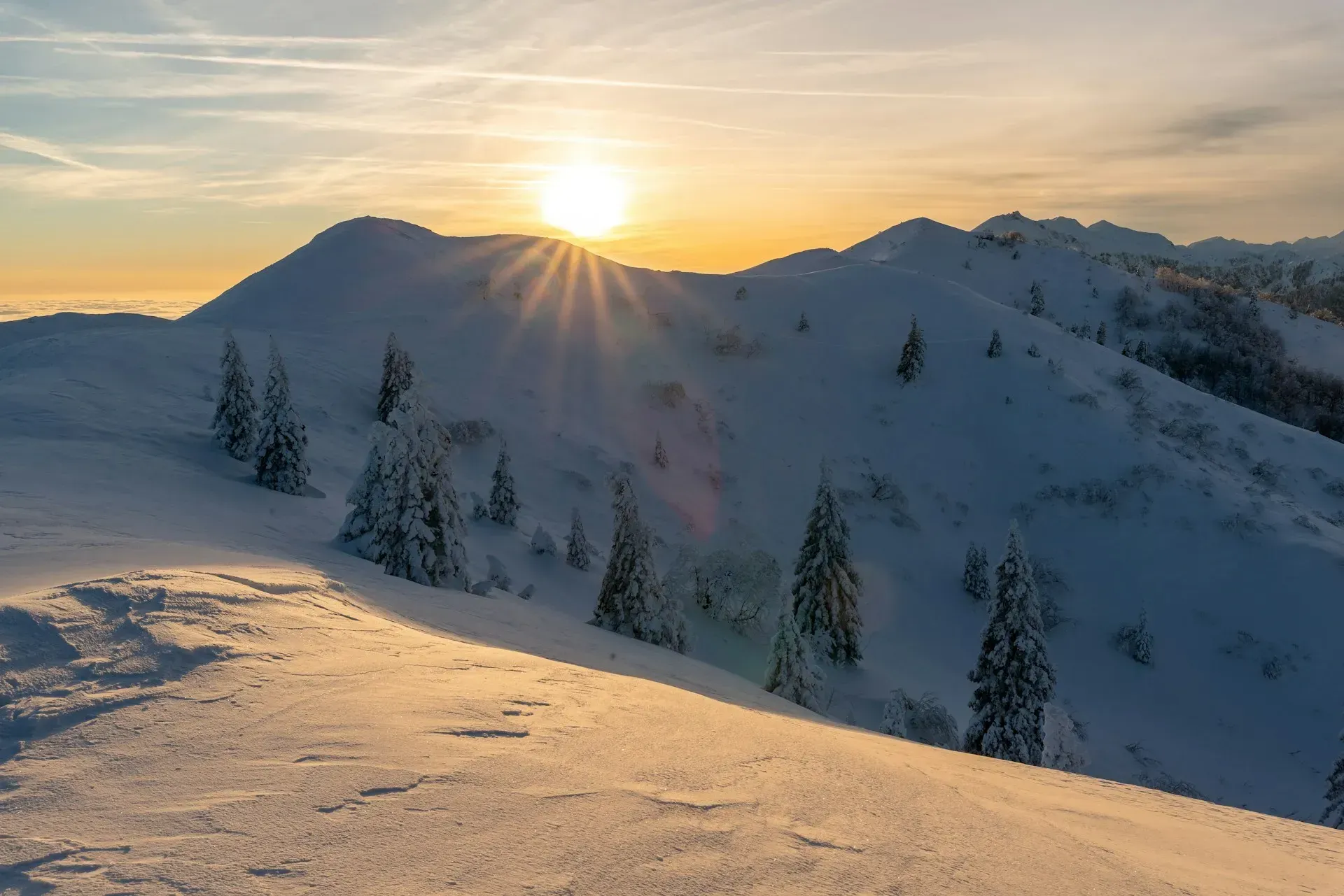 Snowy mountain range at sunrise, sunburst effect. Evergreen trees dot the slopes, golden light.