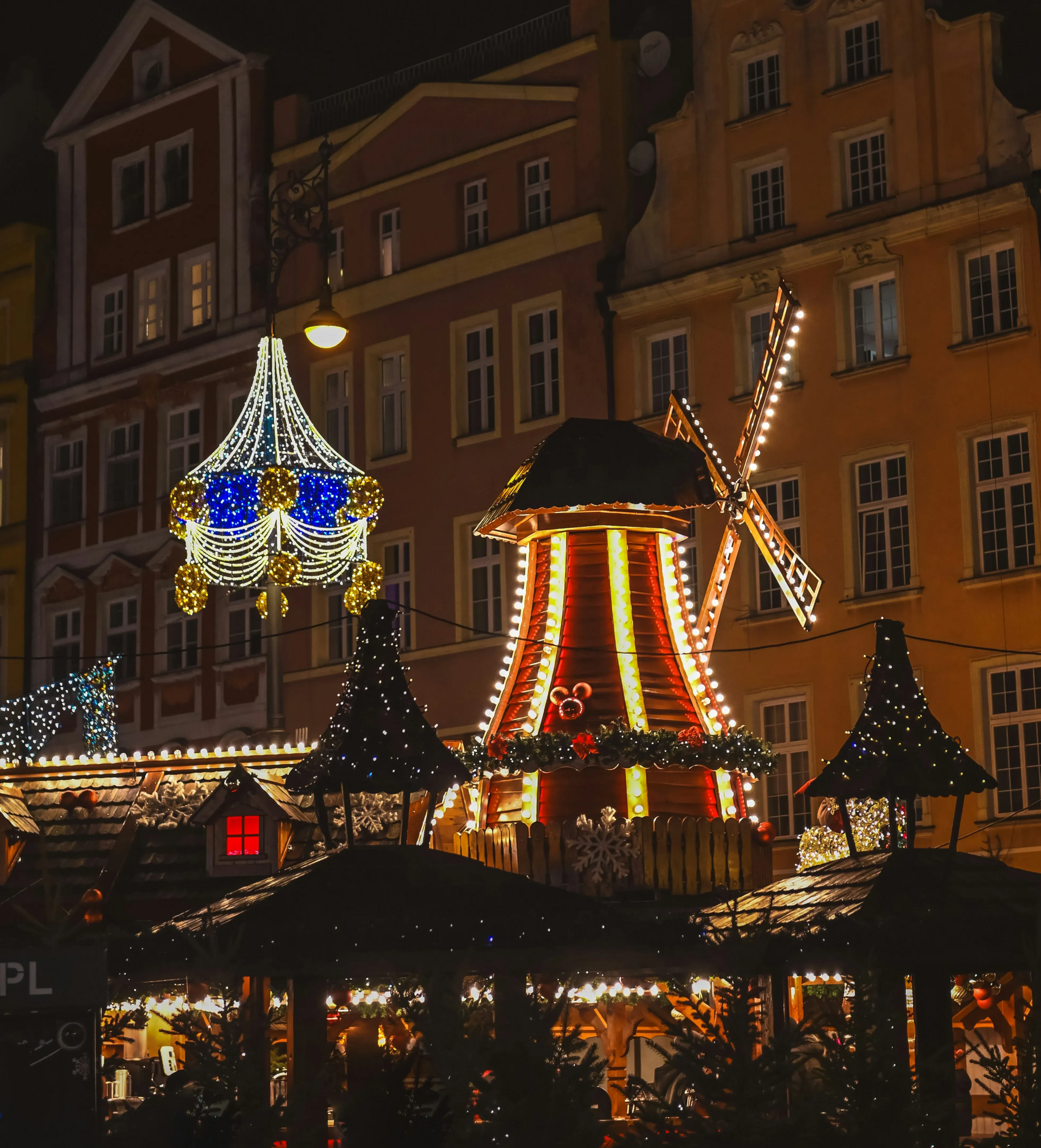 Nocny widok na jarmark bożonarodzeniowy ze świątecznymi światłami, wiatrakiem i dekoracyjną fasadą domu na tle ceglanych budynków.