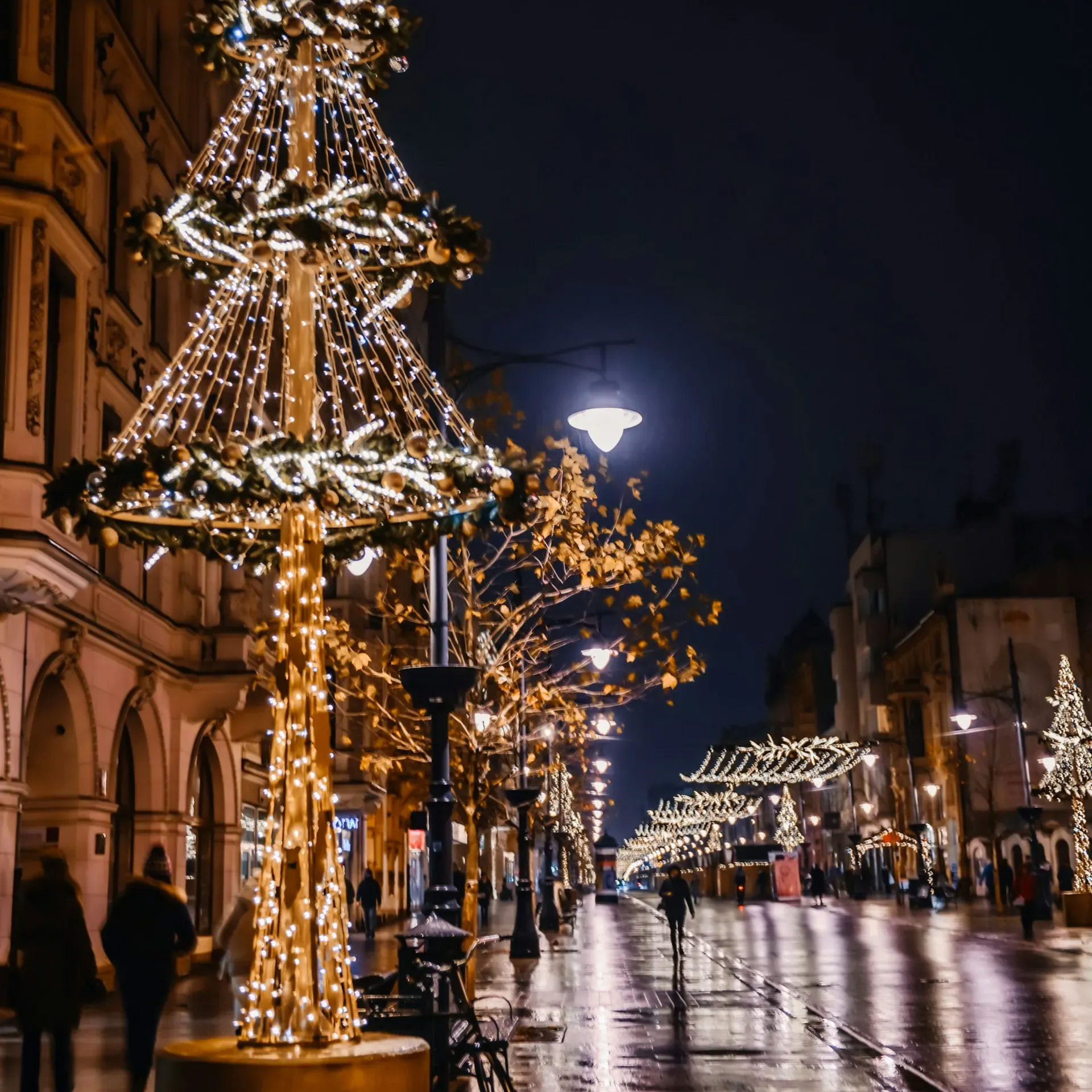 Świąteczne iluminacje oświetlają mokrą ulicę nocą. Ludzie przechodzą obok udekorowanych drzew i budynków.