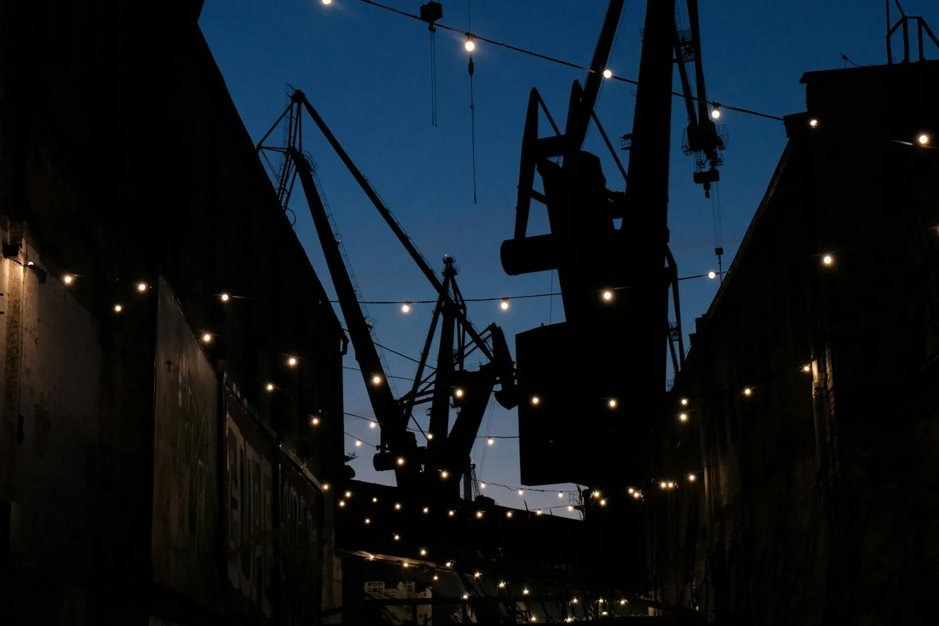 Silhouette of harbor cranes and buildings at dusk, with string lights overhead.