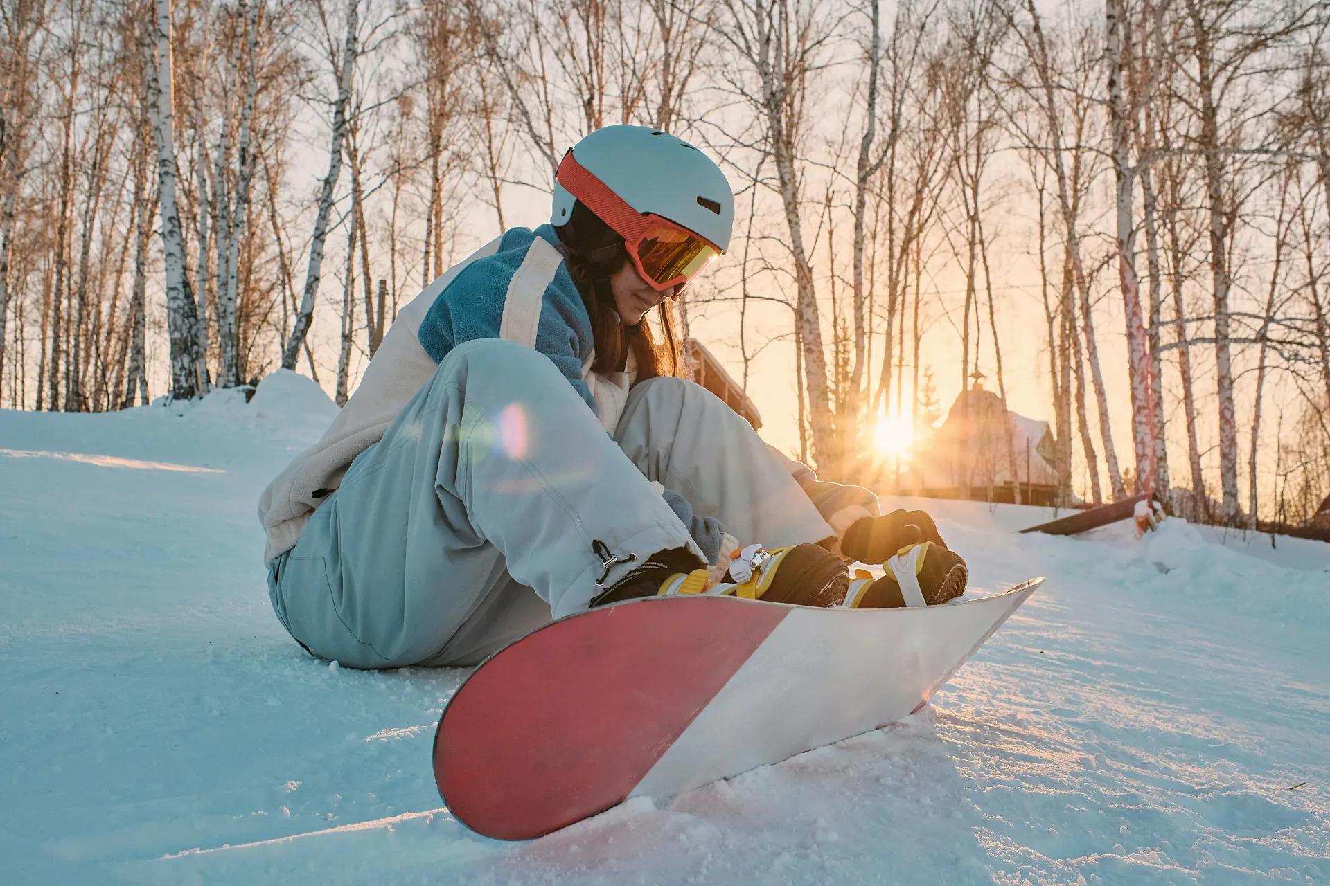 Osoba siedząca na śniegu, zakładająca but snowboardowy. Zachód słońca za drzewami.