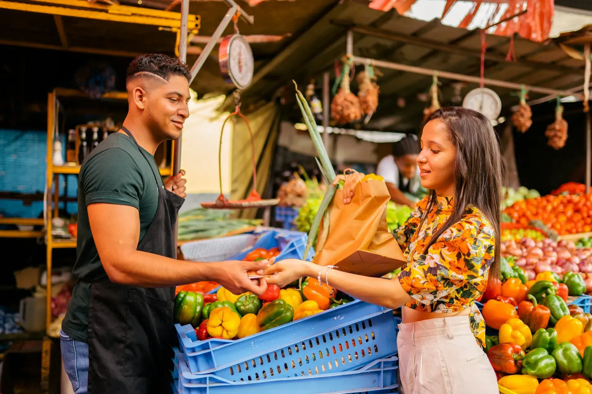 A vendor hands a customer a bag of produce at an outdoor market stall. Colorful vegetables are in crates.