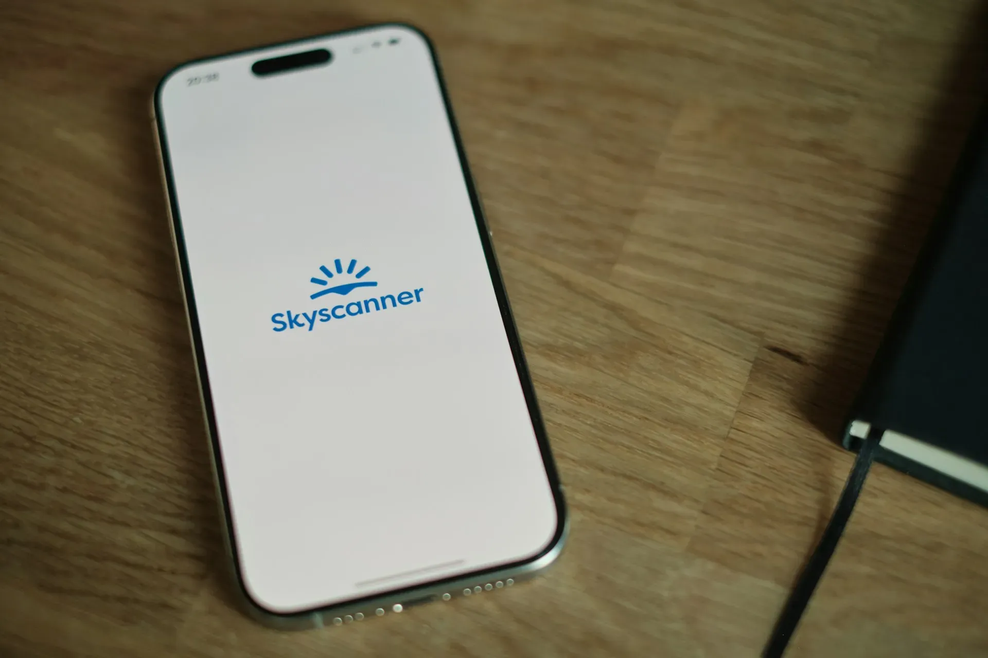 Smartphone displaying the Skyscanner logo on a wooden surface next to a notebook.
