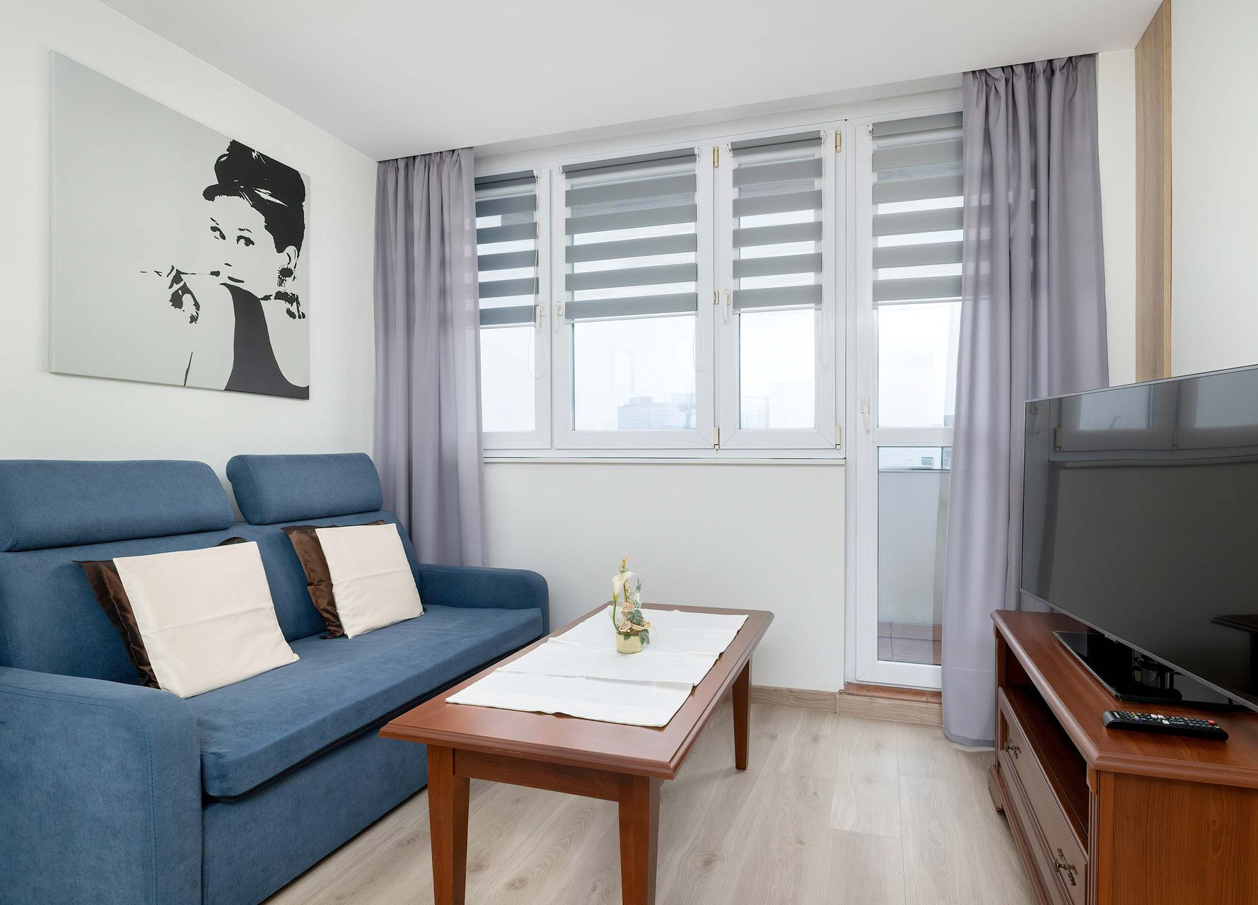 Niebieska kanapa, telewizor i stolik kawowy w małym salonie z balkonem. Dzieło Audrey Hepburn.