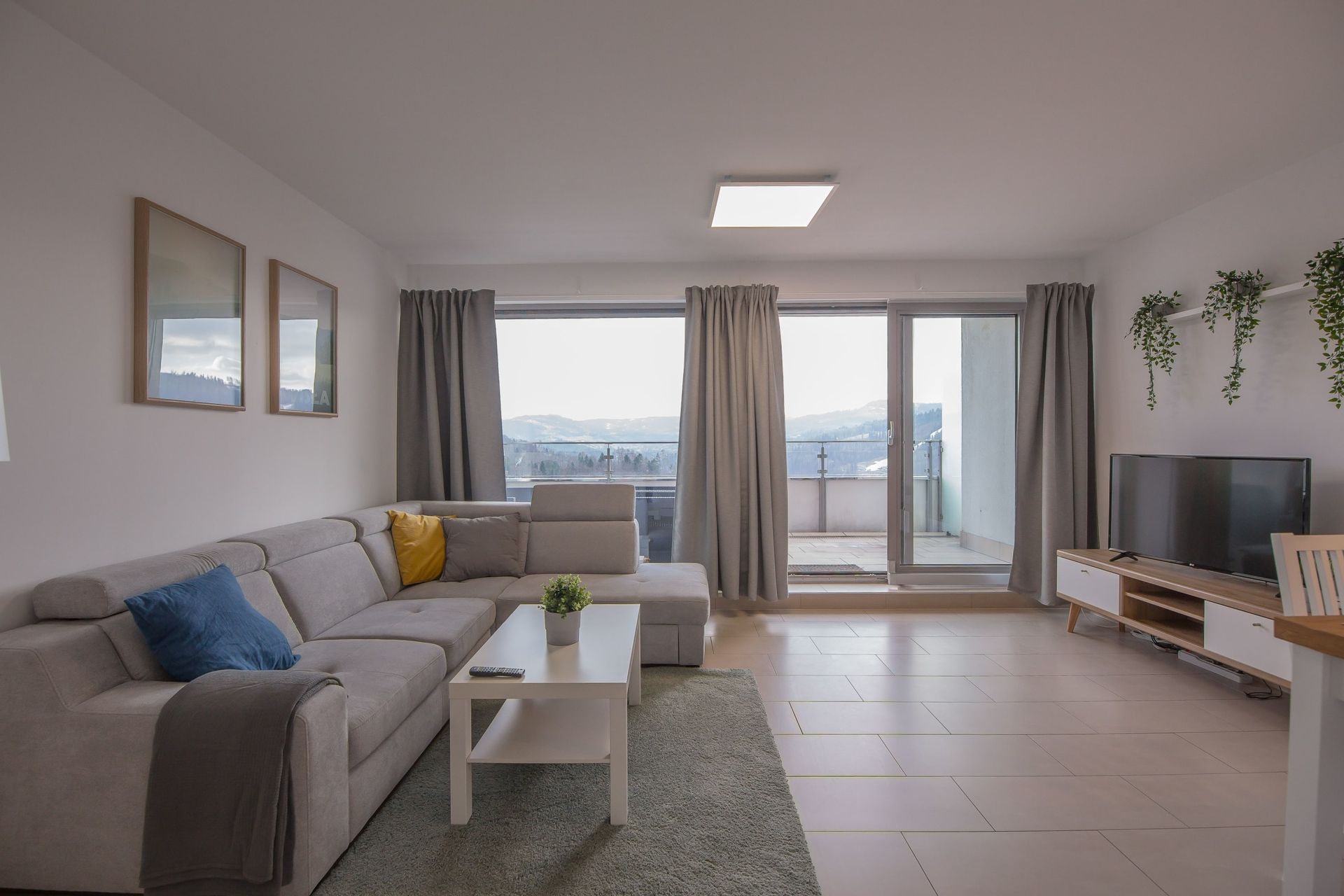 Nowoczesny salon z szarą narożną sofą, stolikiem kawowym, dywanem, szafką pod telewizor i drzwiami balkonowymi z szarymi zasłonami.