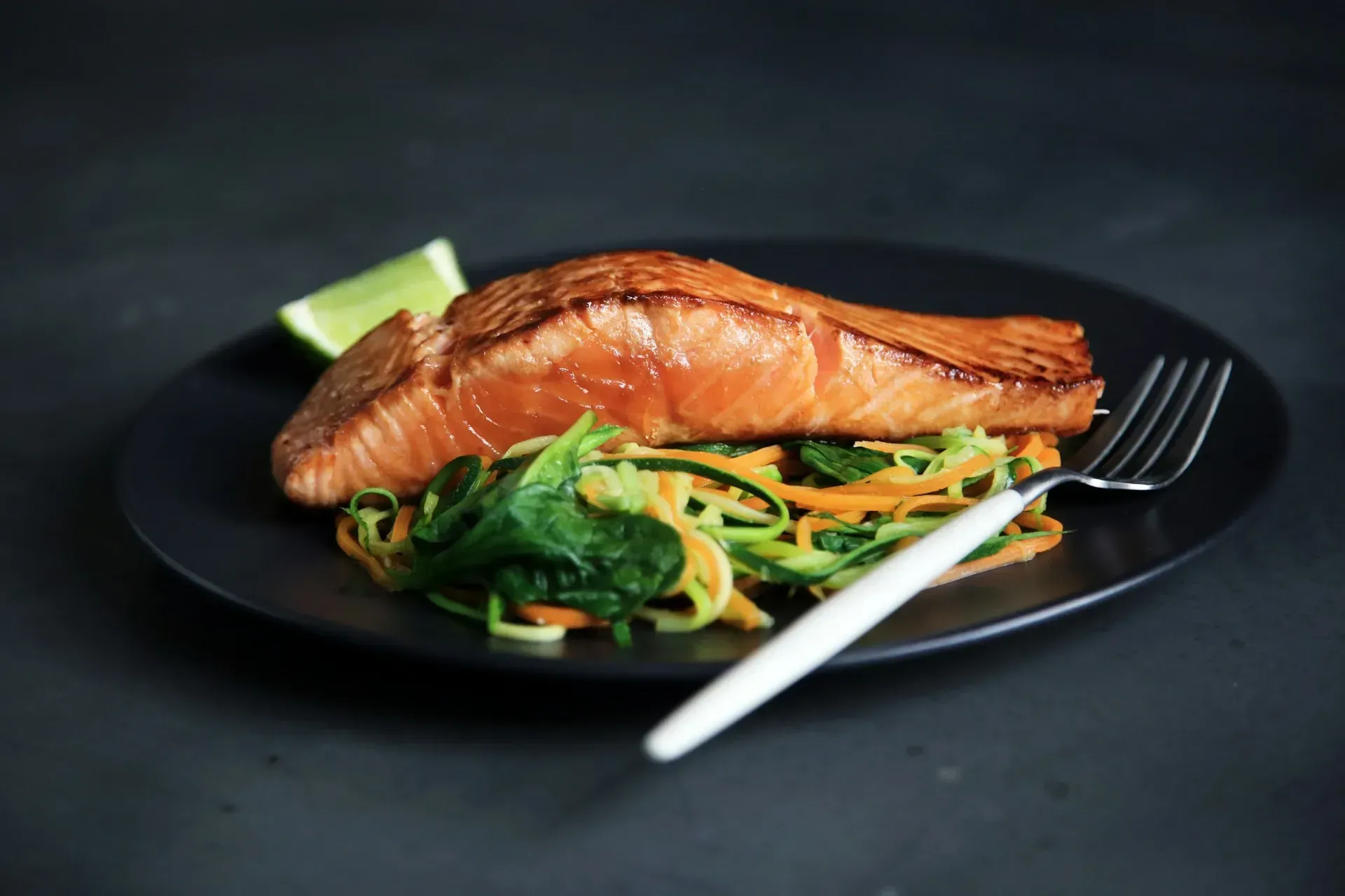 Pan-seared salmon fillet on a bed of vegetables, with a lime wedge on a black plate.