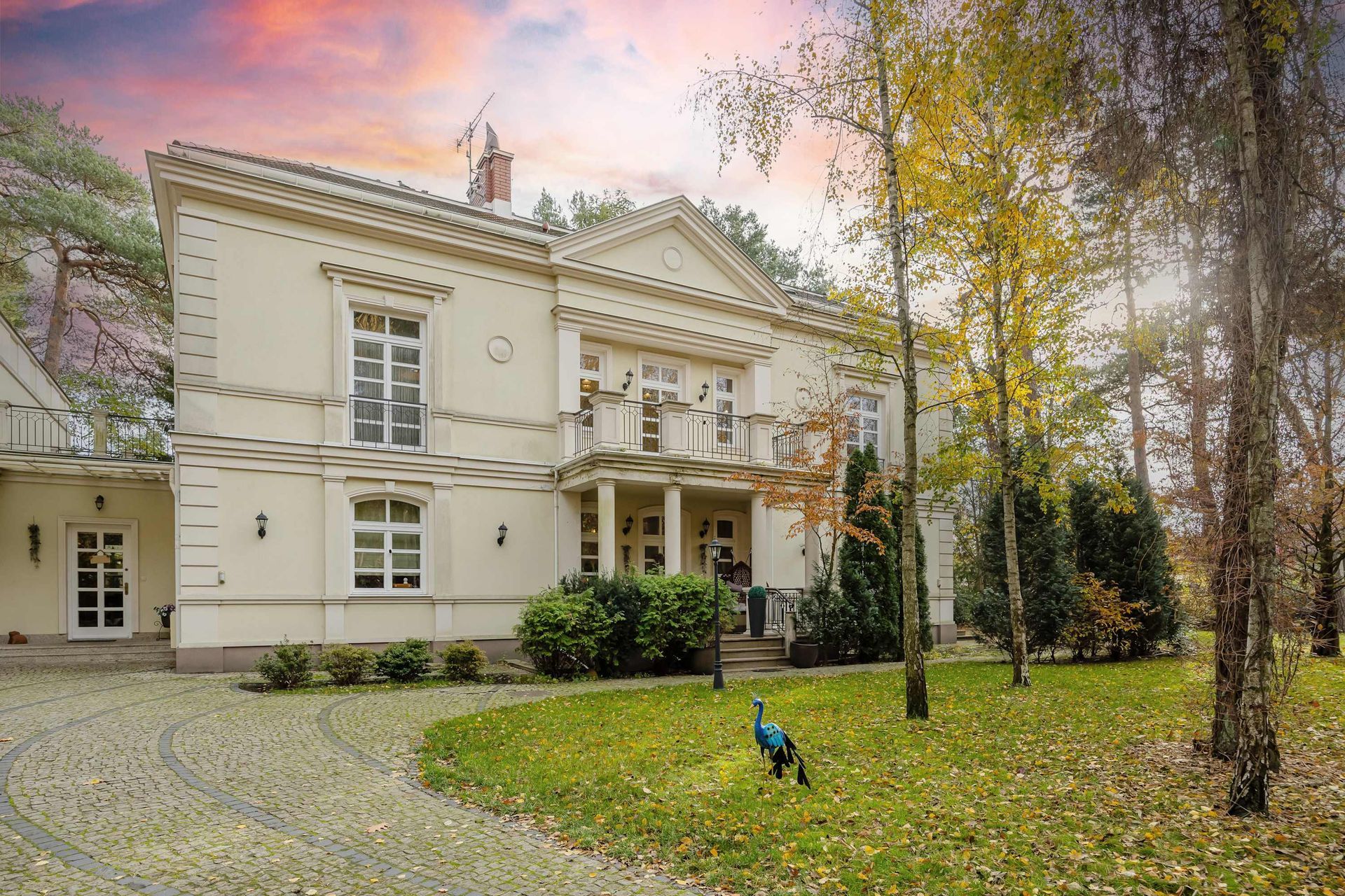 Elegancka, biała rezydencja z portykiem, balkonem i podjazdem, otoczona drzewami i pawiem na trawniku.