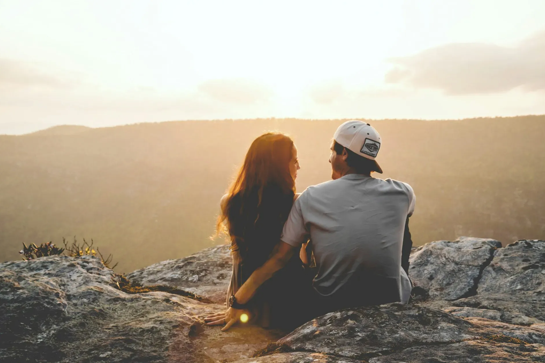 Couple sitting on a rocky cliff, looking at each other as the sun sets over distant mountains.