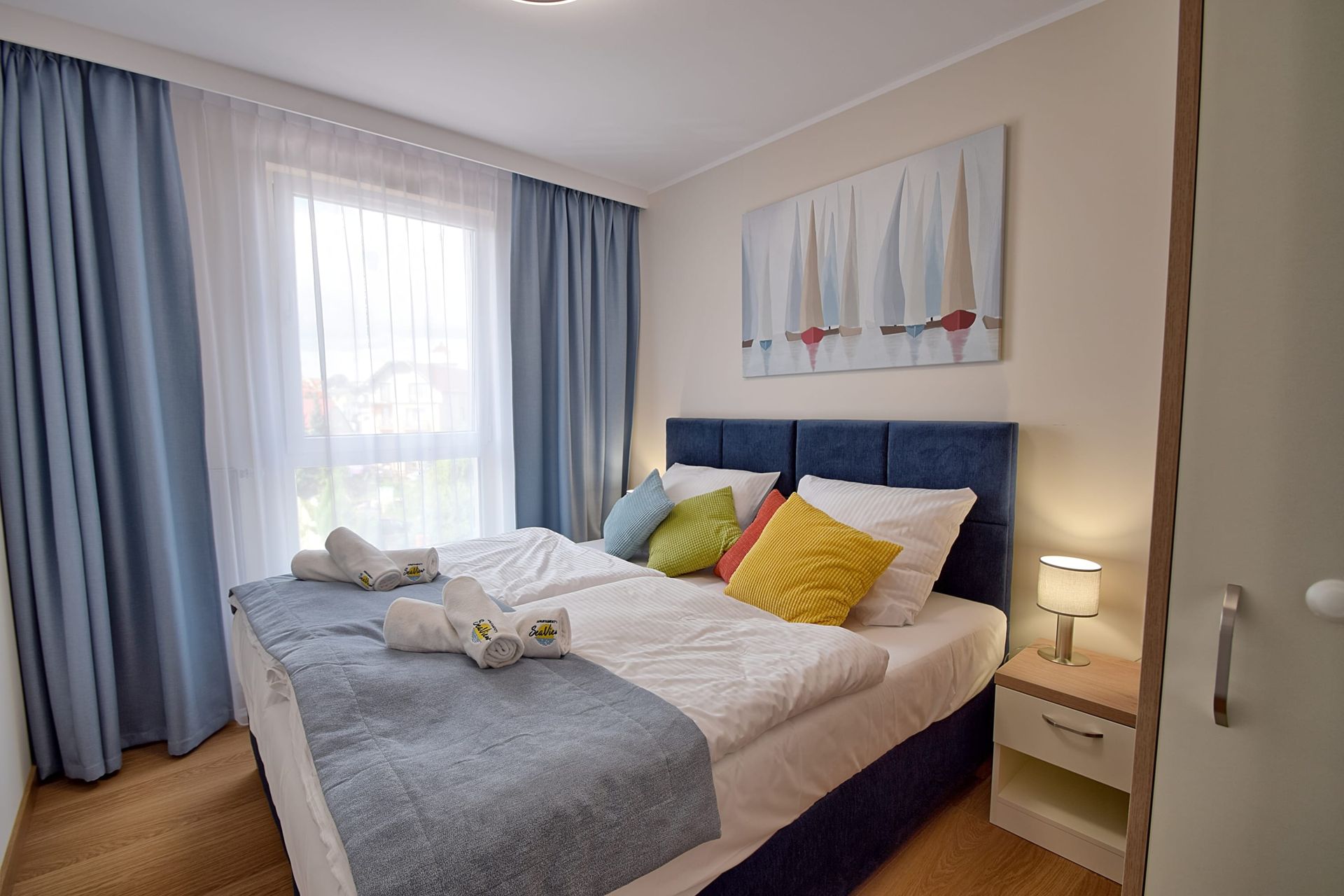 Pokój hotelowy z łóżkiem z niebieskim zagłówkiem, białą pościelą, kolorowymi poduszkami, niebieskimi zasłonami i malowidłem ściennym.