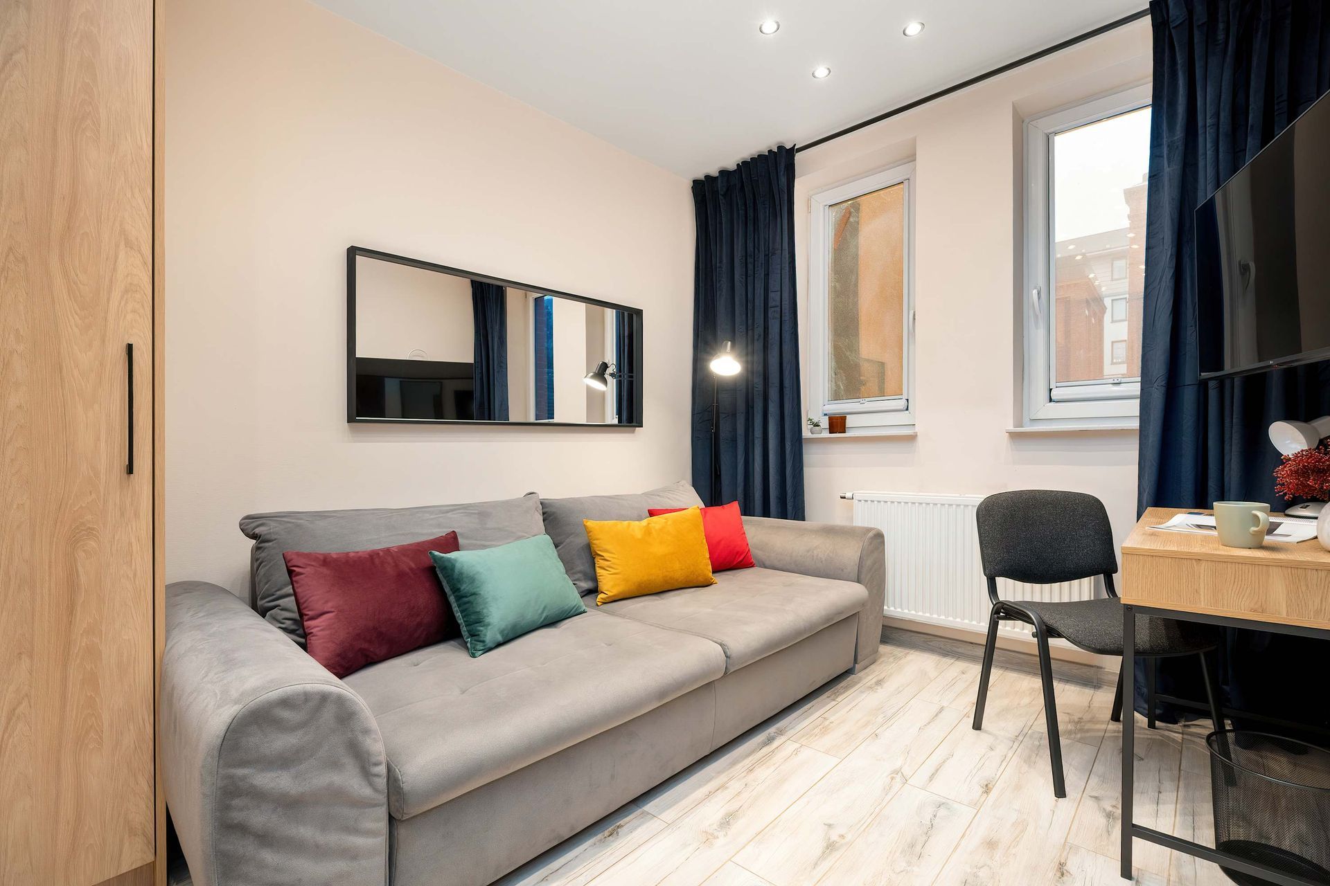 Kompaktowe studio z szarą sofą, kolorowymi poduszkami, biurkiem, krzesłem i dużymi oknami z ciemnymi zasłonami.