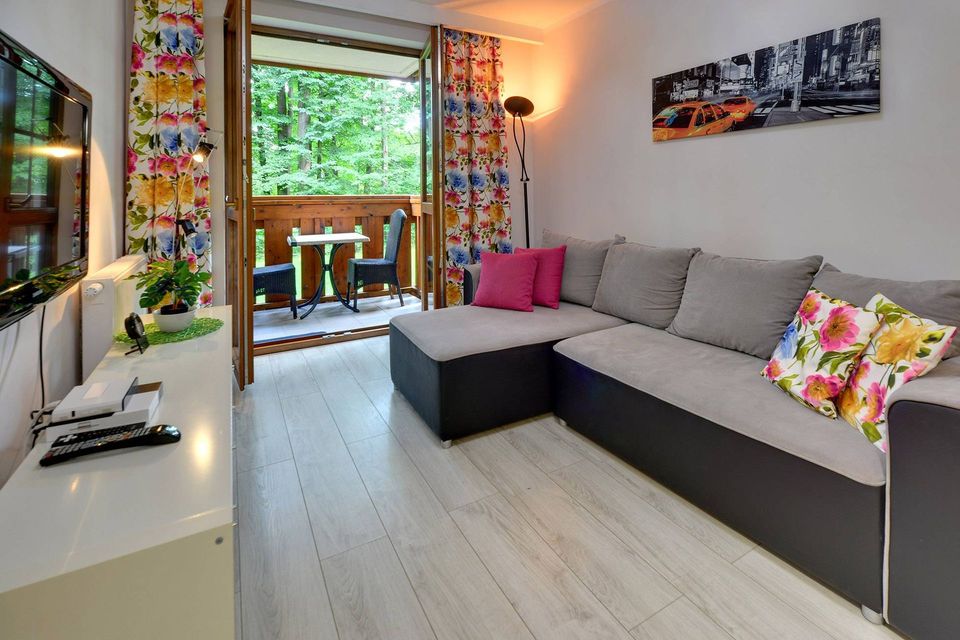 Nowoczesny salon z szarą sofą narożną, kolorowymi poduszkami i przesuwnymi drzwiami prowadzącymi na balkon pełen zieleni.