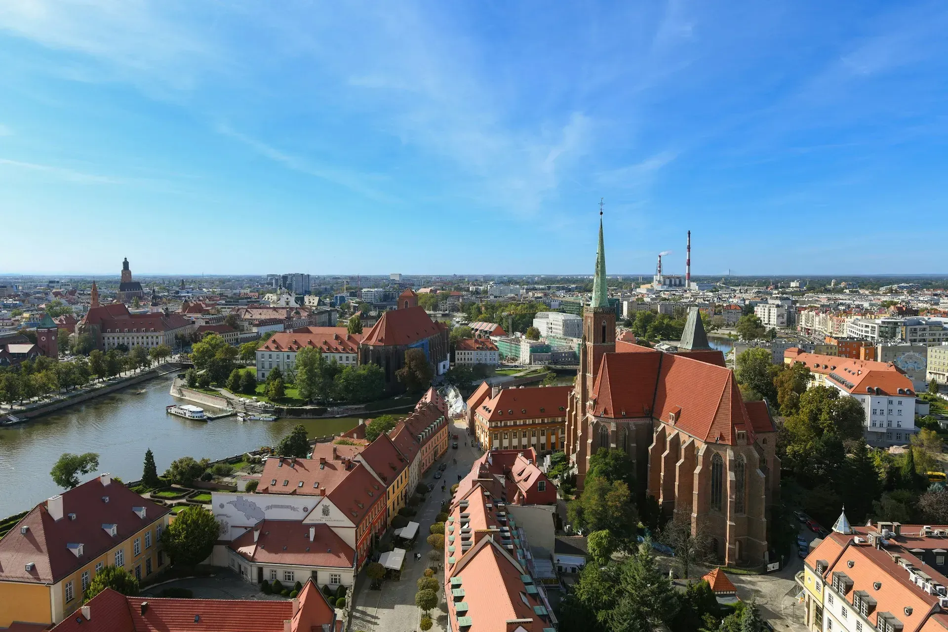 Aerial view of a city with red-roofed buildings, a river, and a tall church spire under a blue sky.