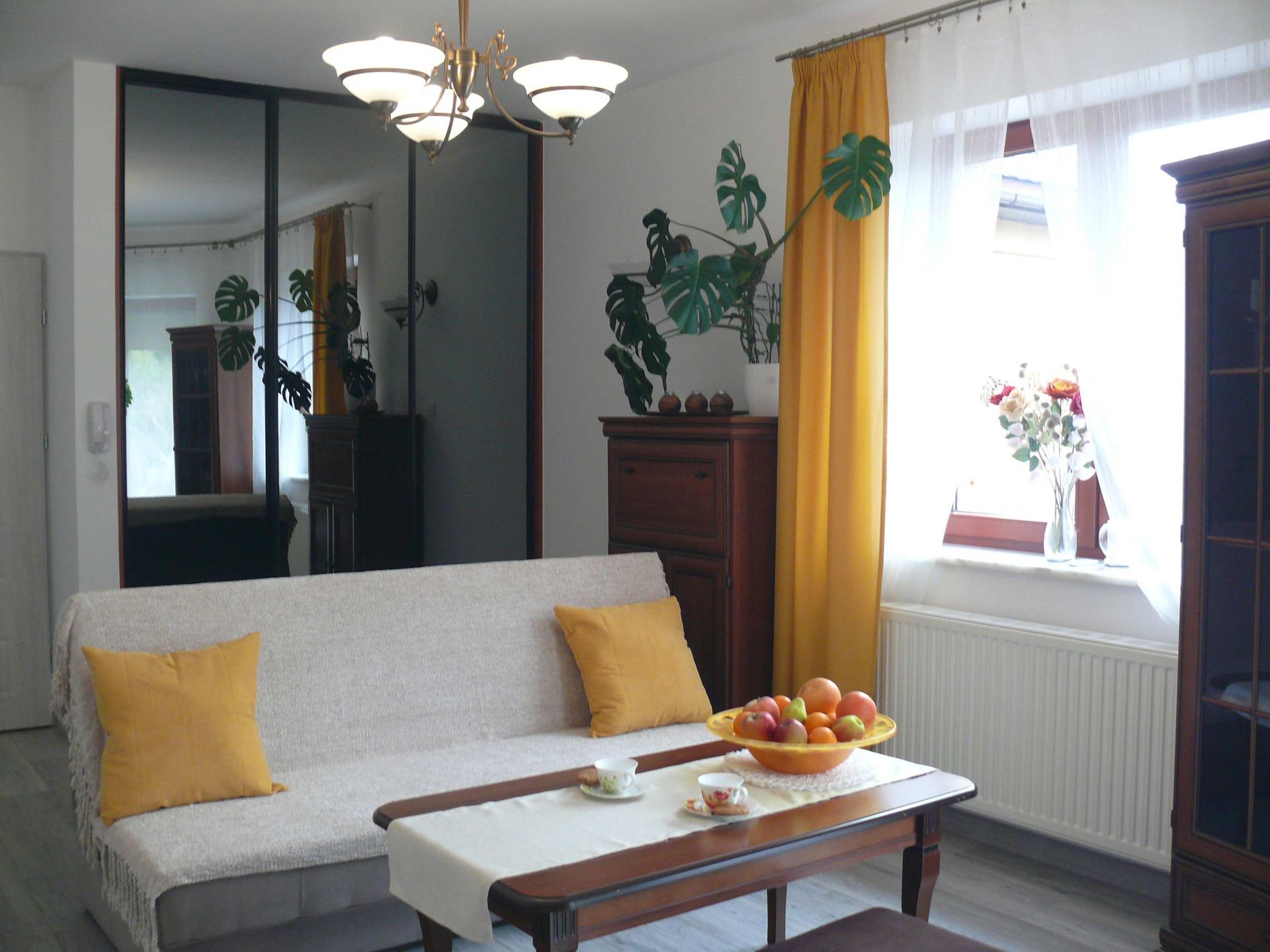 Salon z sofą, stołem, komodą, lustrem i żółtymi zasłonami.