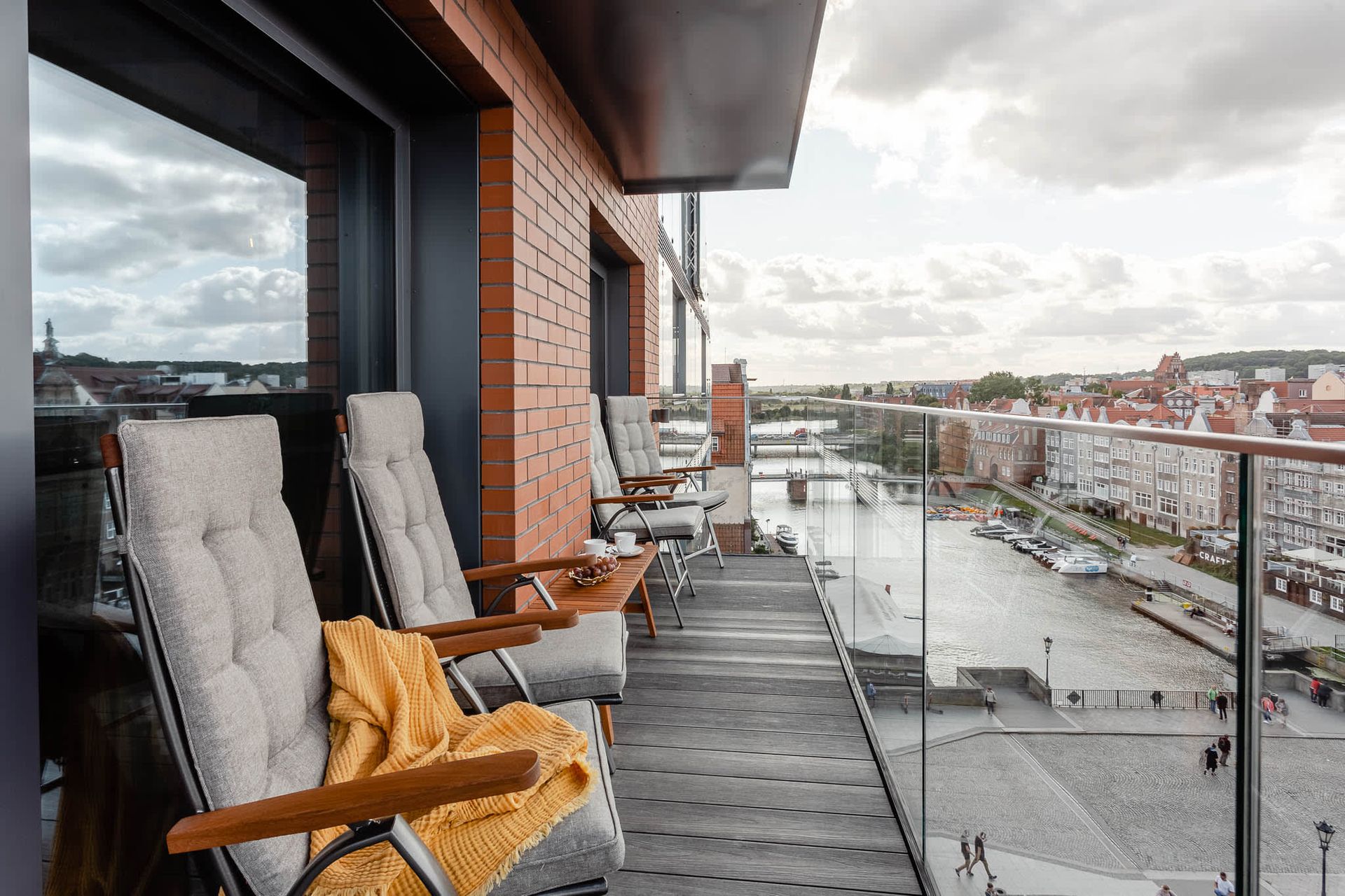 Balkon z drewnianym tarasem, krzesłami i widokiem na port. Budynek Deo Plaza z cegły, zachmurzone niebo, neutralne kolory.