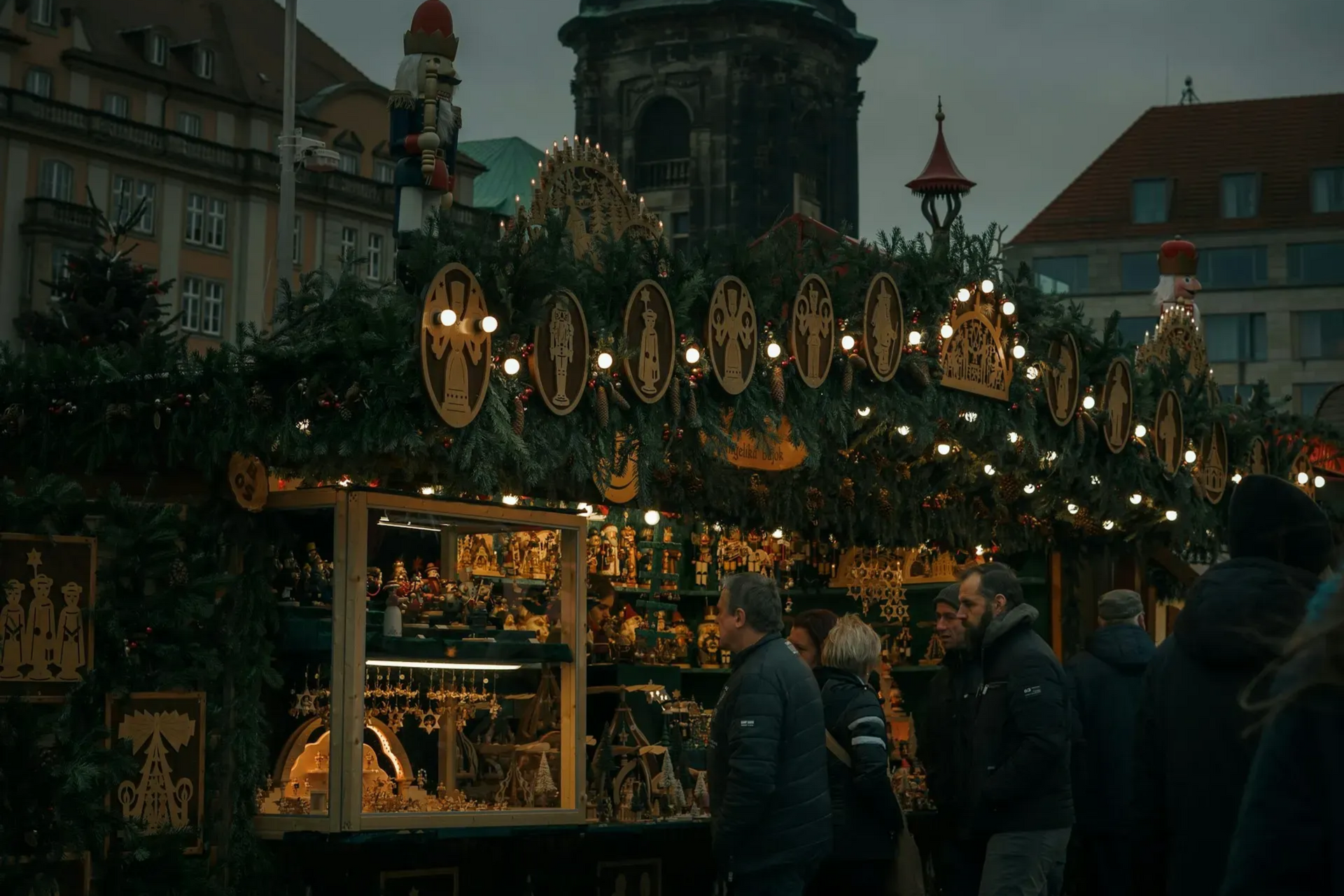 Christmas market stall with wooden crafts, adorned with lights and greenery; people browsing.