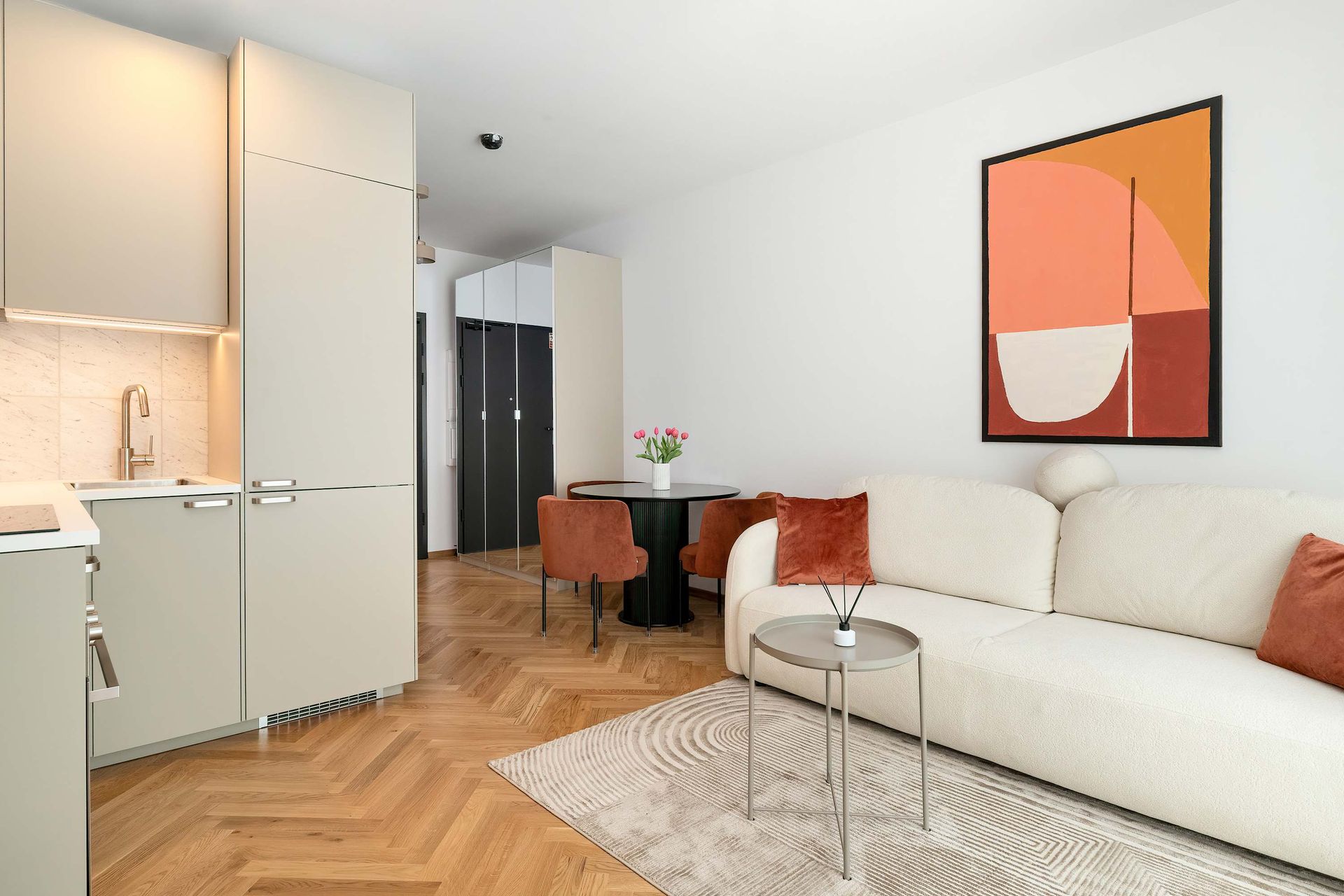 Nowoczesny salon z beżową sofą, małym stolikiem kawowym, kącikiem jadalnym i aneksem kuchennym z jasną drewnianą podłogą.