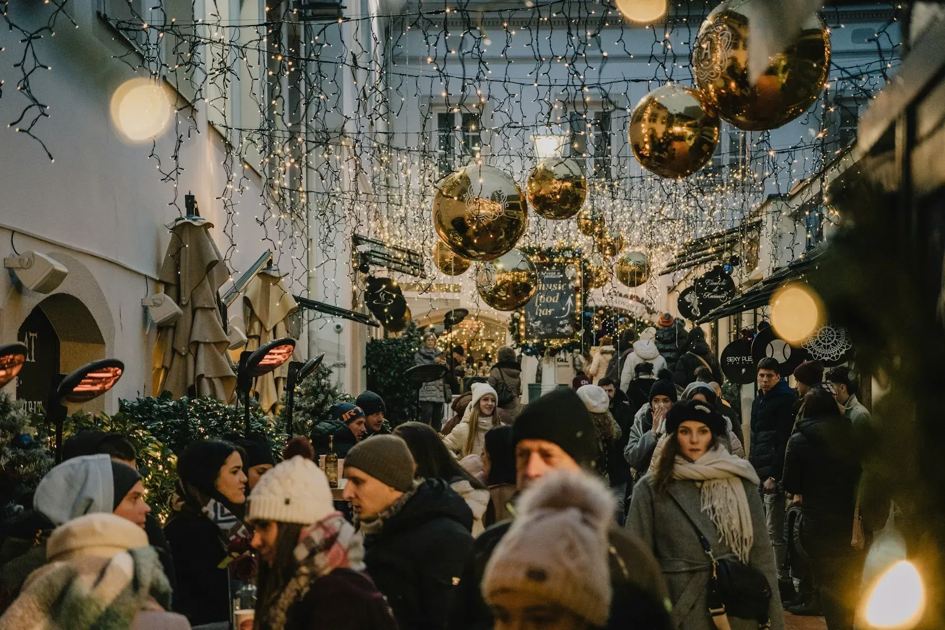 Crowd of people walking under Christmas lights and gold ornaments in a festive outdoor alley.