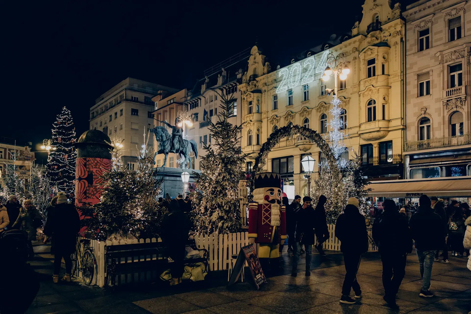 Night scene of a European square with Christmas decorations, lit buildings, and people.