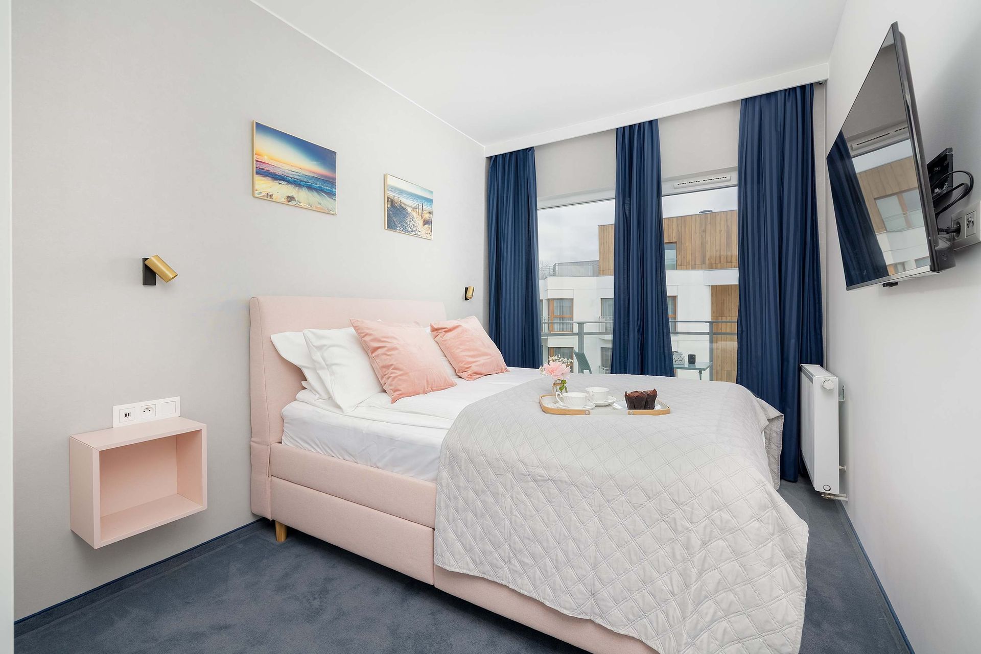 Sypialnia z różowym łóżkiem, niebieskimi zasłonami i zamontowanym telewizorem.